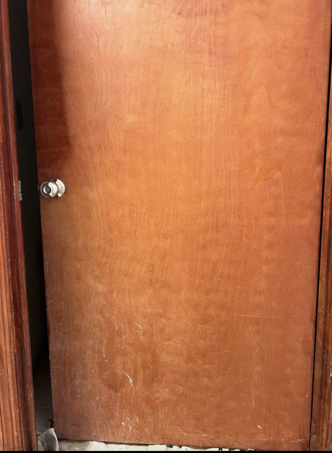 A closed wooden door with a round metallic doorknob, partially open, revealing a dark interior. The door has a reddish-brown color and visible wood grain.