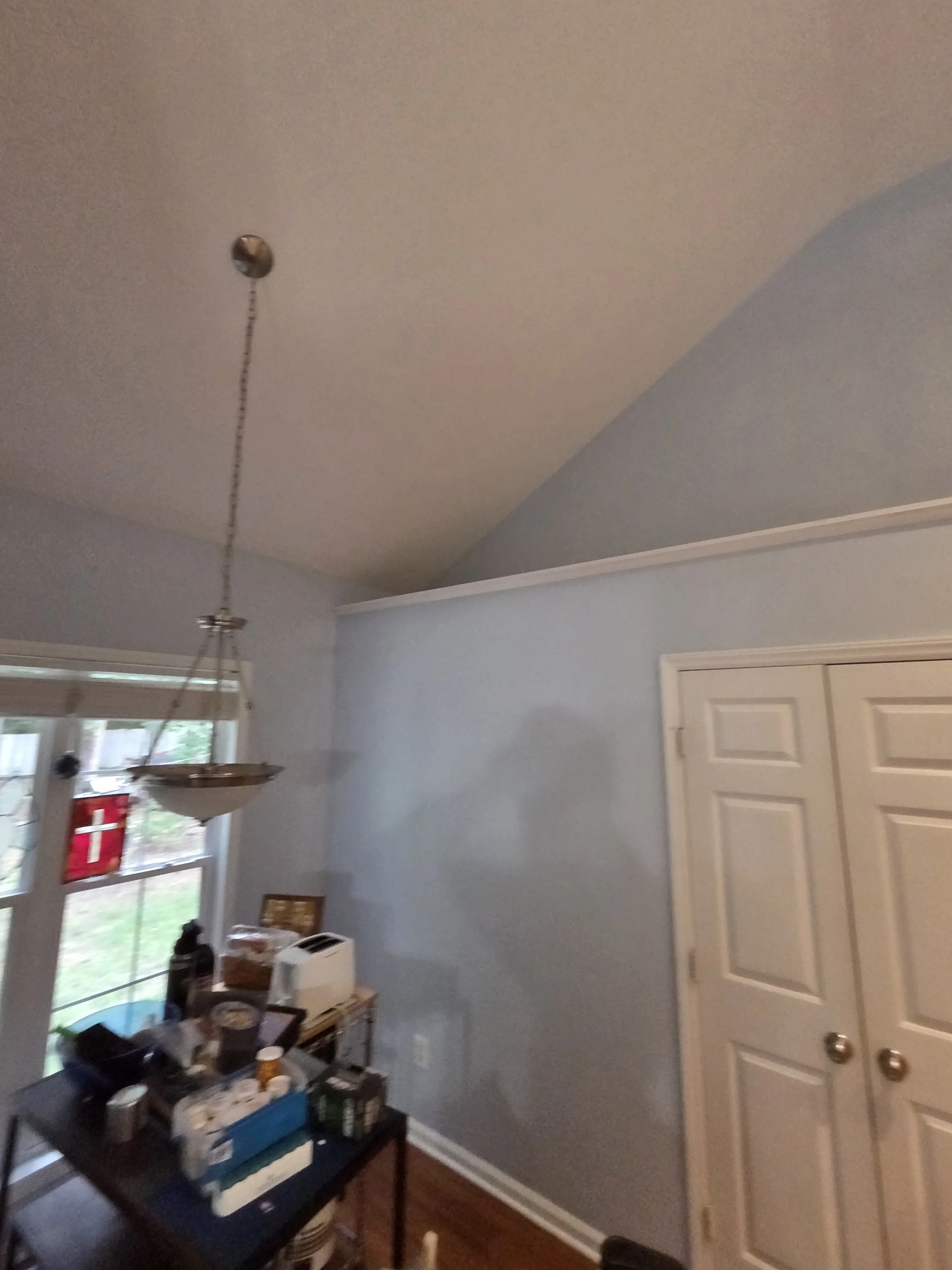 A room with gray walls and a white ceiling, featuring a plastic closet door, a window with blinds, a shelf with various objects, and a hanging light fixture.