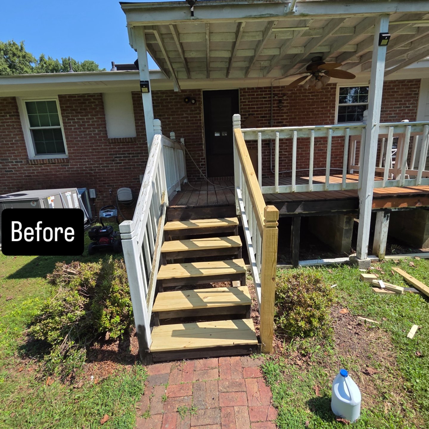 Backyard deck construction with wooden stairs, partially built railings, and a brick house in the background. Tools and construction materials are on the ground and an air conditioning unit is visible.