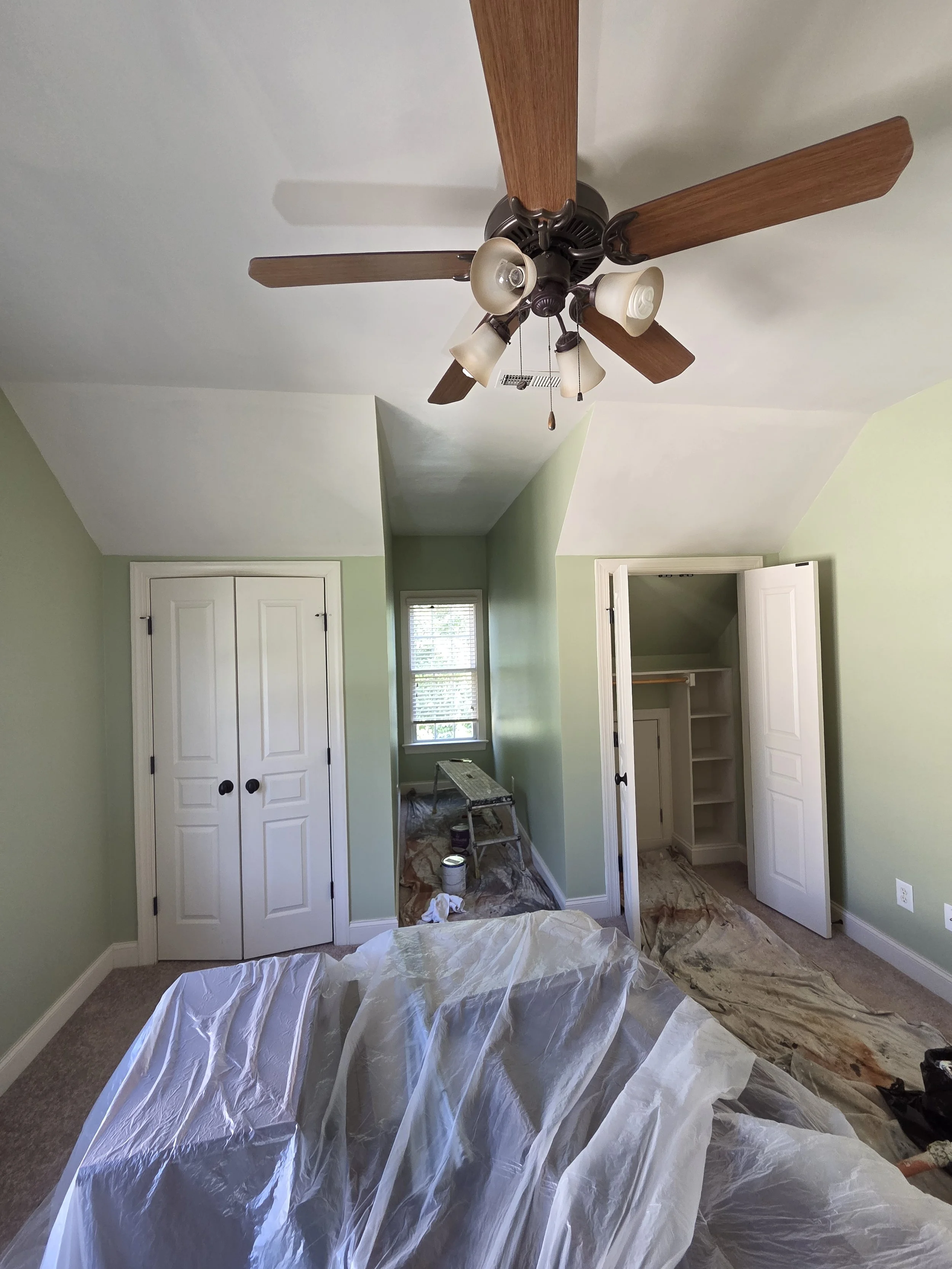 Room undergoing renovation with plastic sheeting cover on the floor, paint supplies, an open closet, a closed double door, and a ceiling fan with lights.