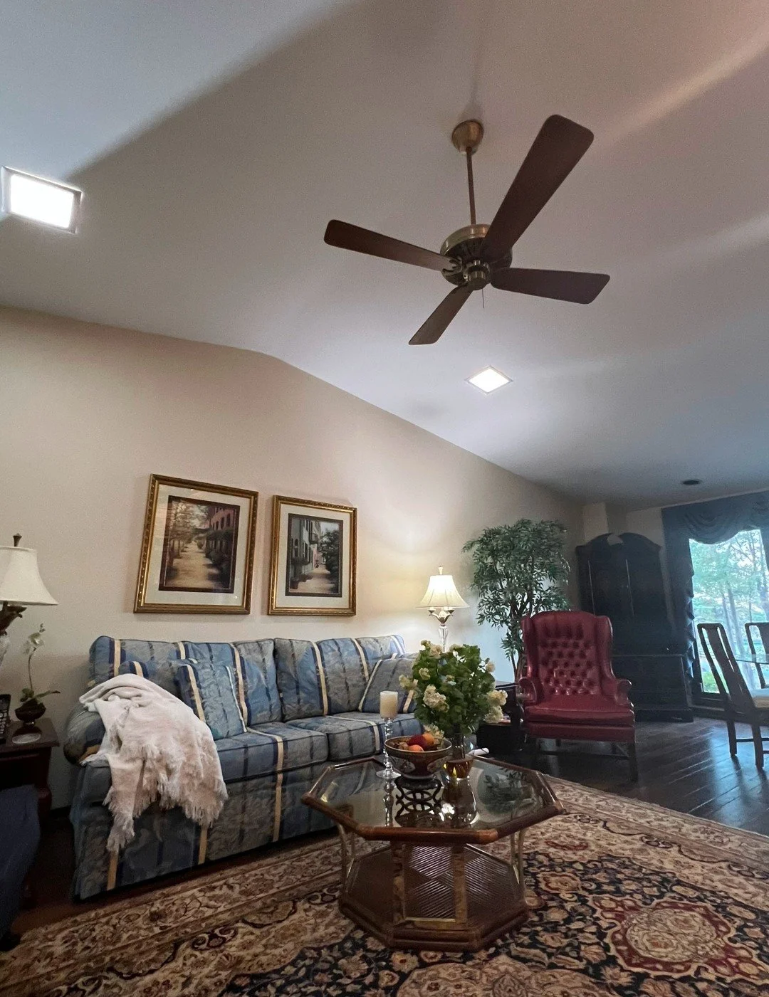 Living room with ceiling fan, framed pictures, sofa with throw blanket, side tables with lamps, glass coffee table with flowers, armchair, and window with curtains.