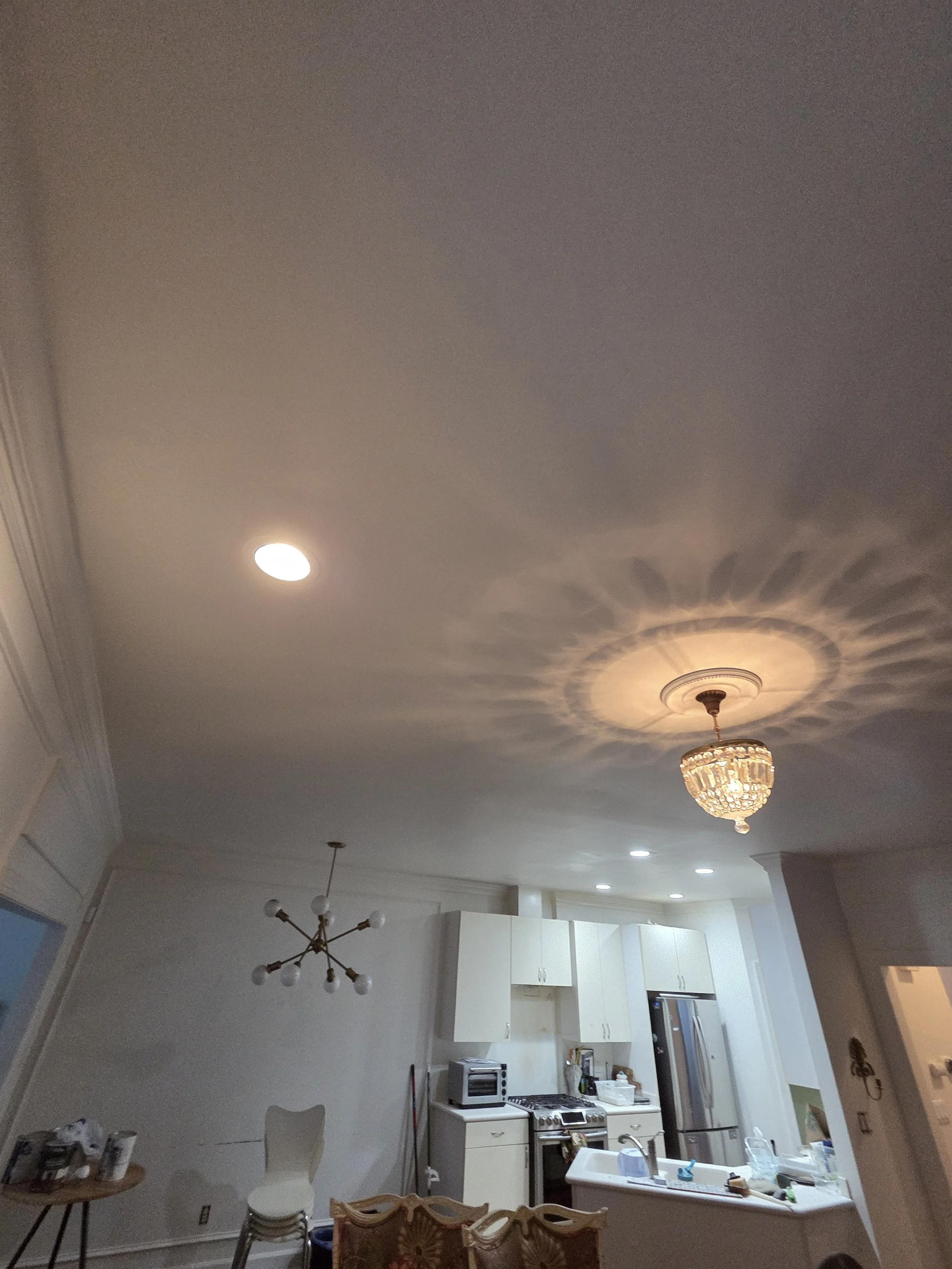 Living room ceiling with recessed lights and decorative chandelier, kitchen with white cabinets, stainless steel appliances, and a kitchen island with a sink, part of a dining area with chairs.