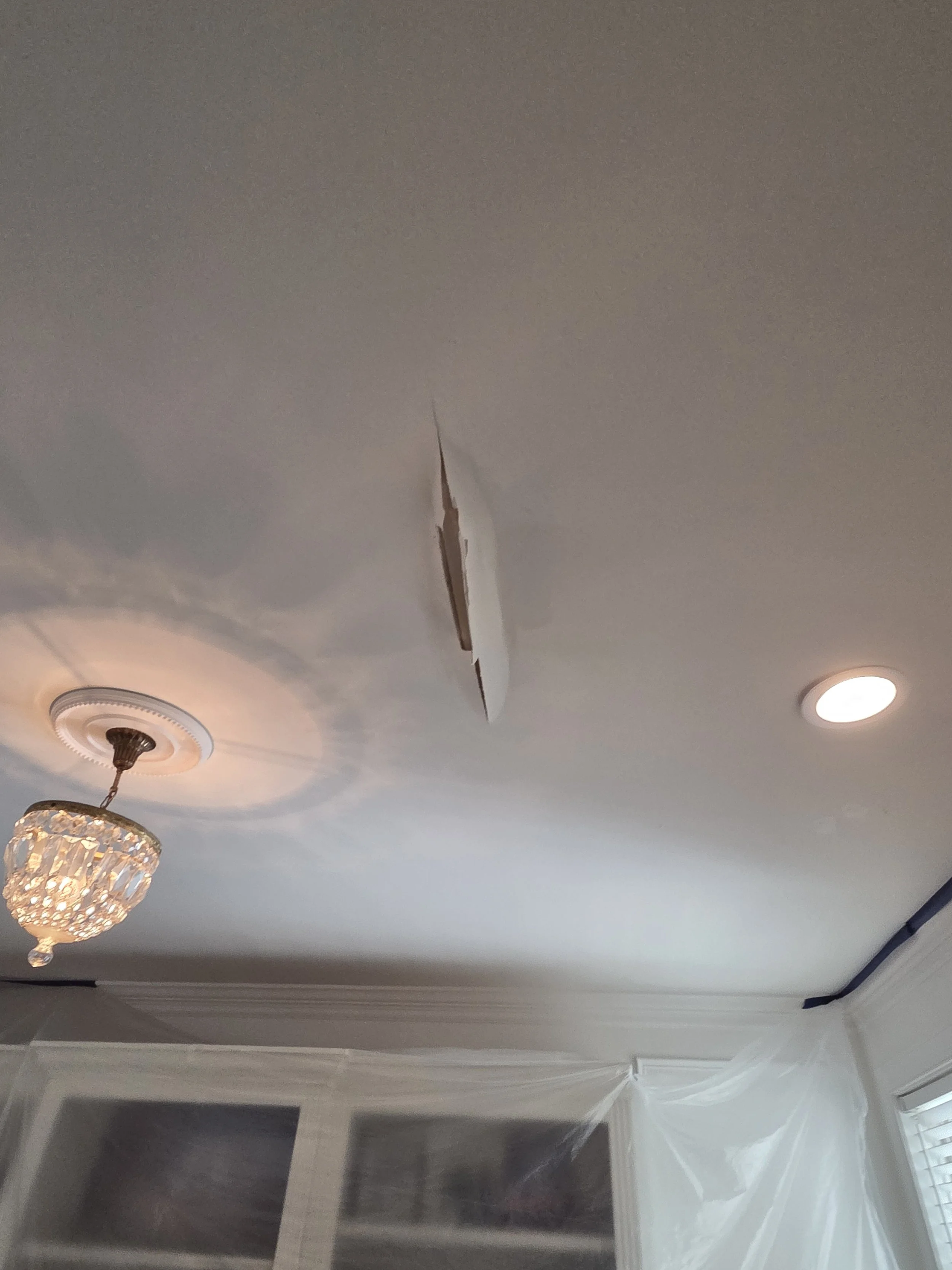 Ceiling with a missing drywall section, a chandelier, a ceiling light, and part of a window with blinds.
