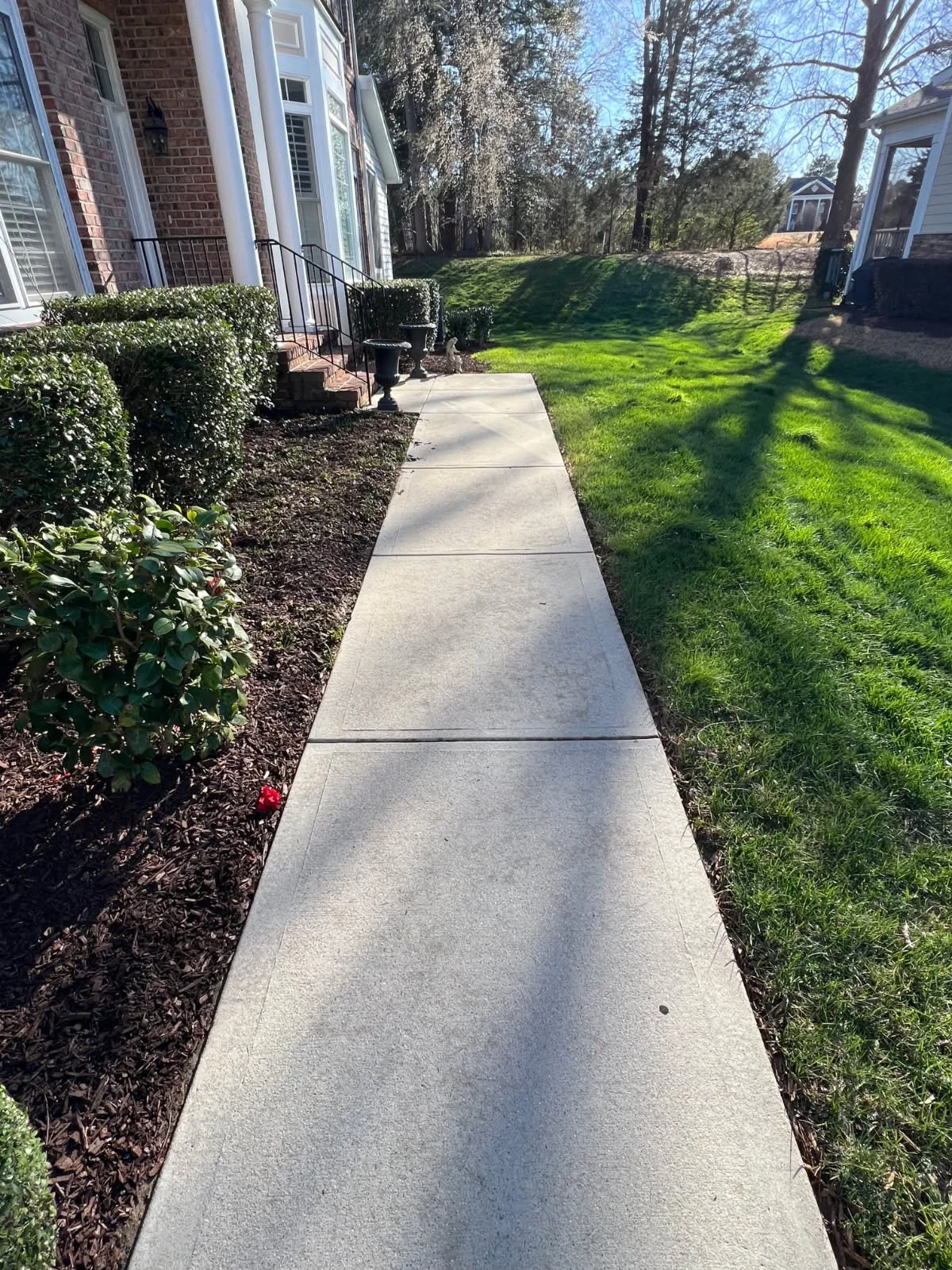 Concrete sidewalk running alongside a house with brick and siding exterior, green bushes on the left, and a grassy yard on the right, with trees and a blue sky in the background.