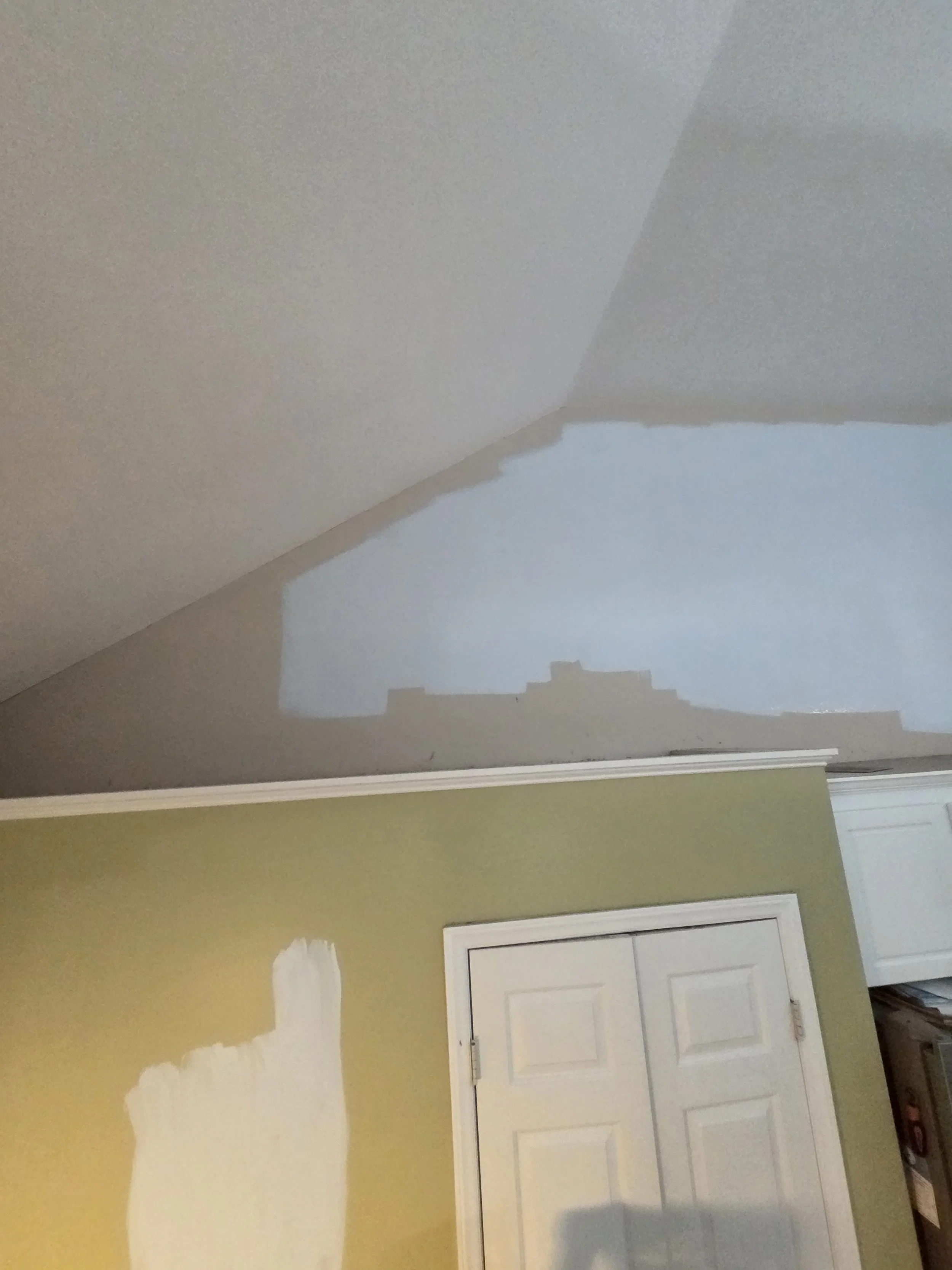 A room undergoing renovation, with drywall patches on a yellow wall, a white door with a four-panel design, and a white window blind visible at the bottom right corner.