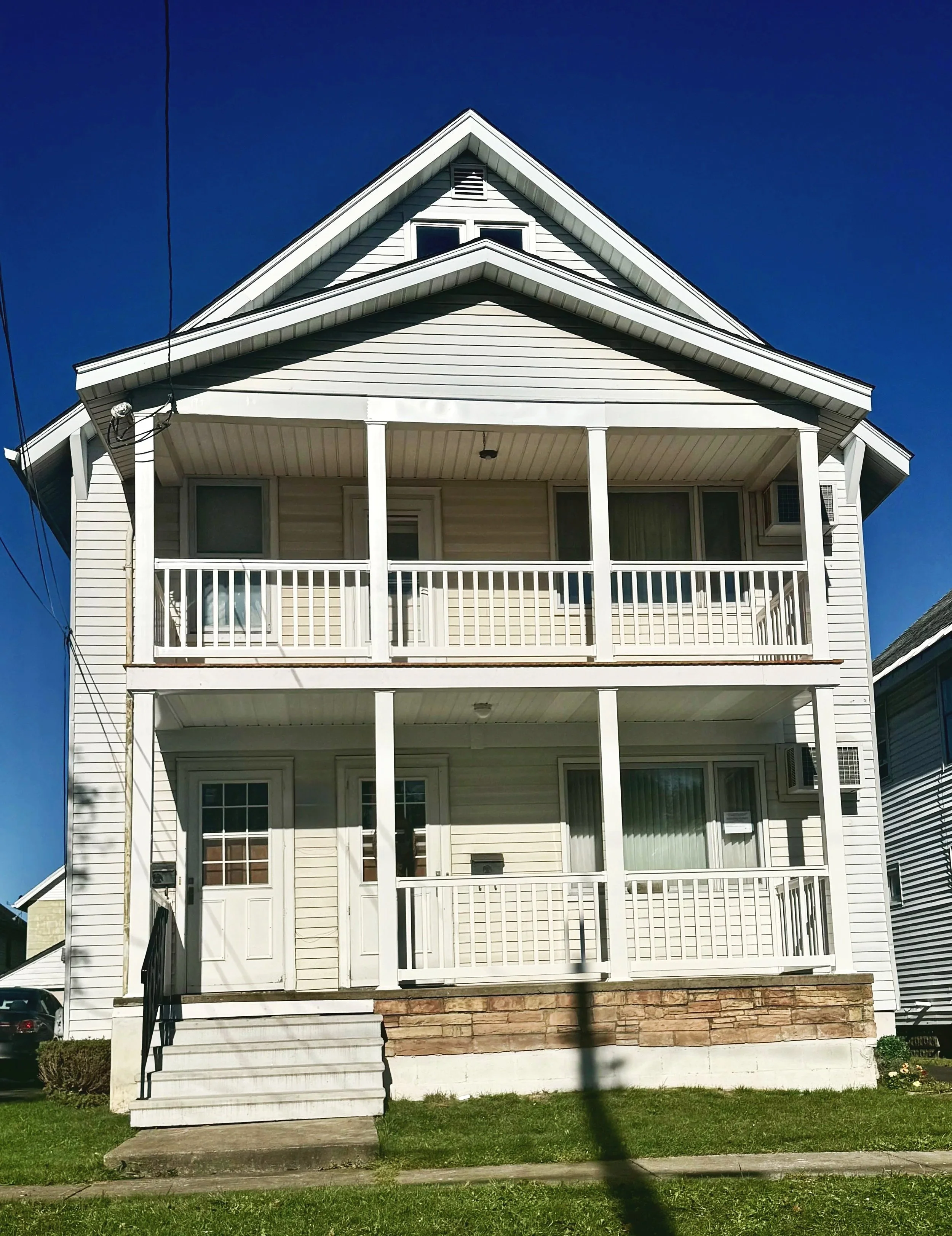 A three-story white house with two front porches, each with white railings and columns. The house has beige siding, a gable roof, and a staircase leading to the front door. The sky is clear and blue. Porch. 2 story.