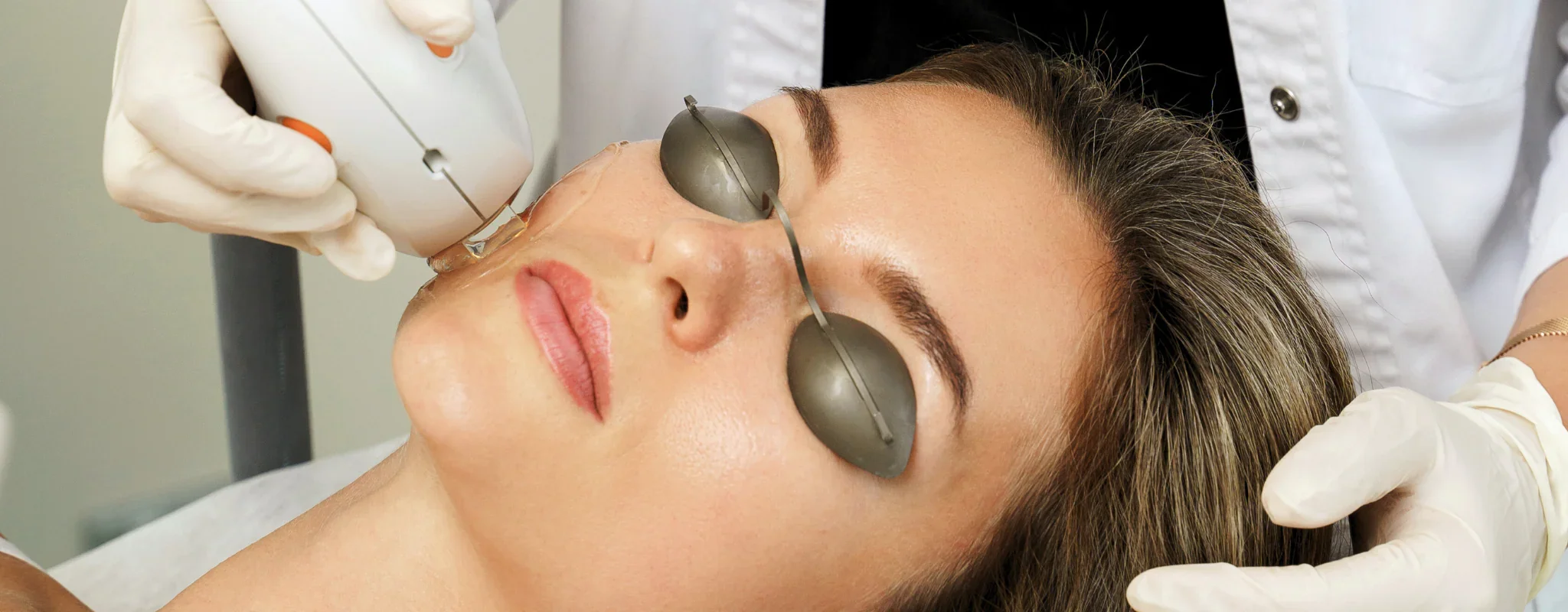 A woman receiving a cosmetic facial treatment with a medical device while wearing protective eye goggles, supervised by a healthcare professional in gloves