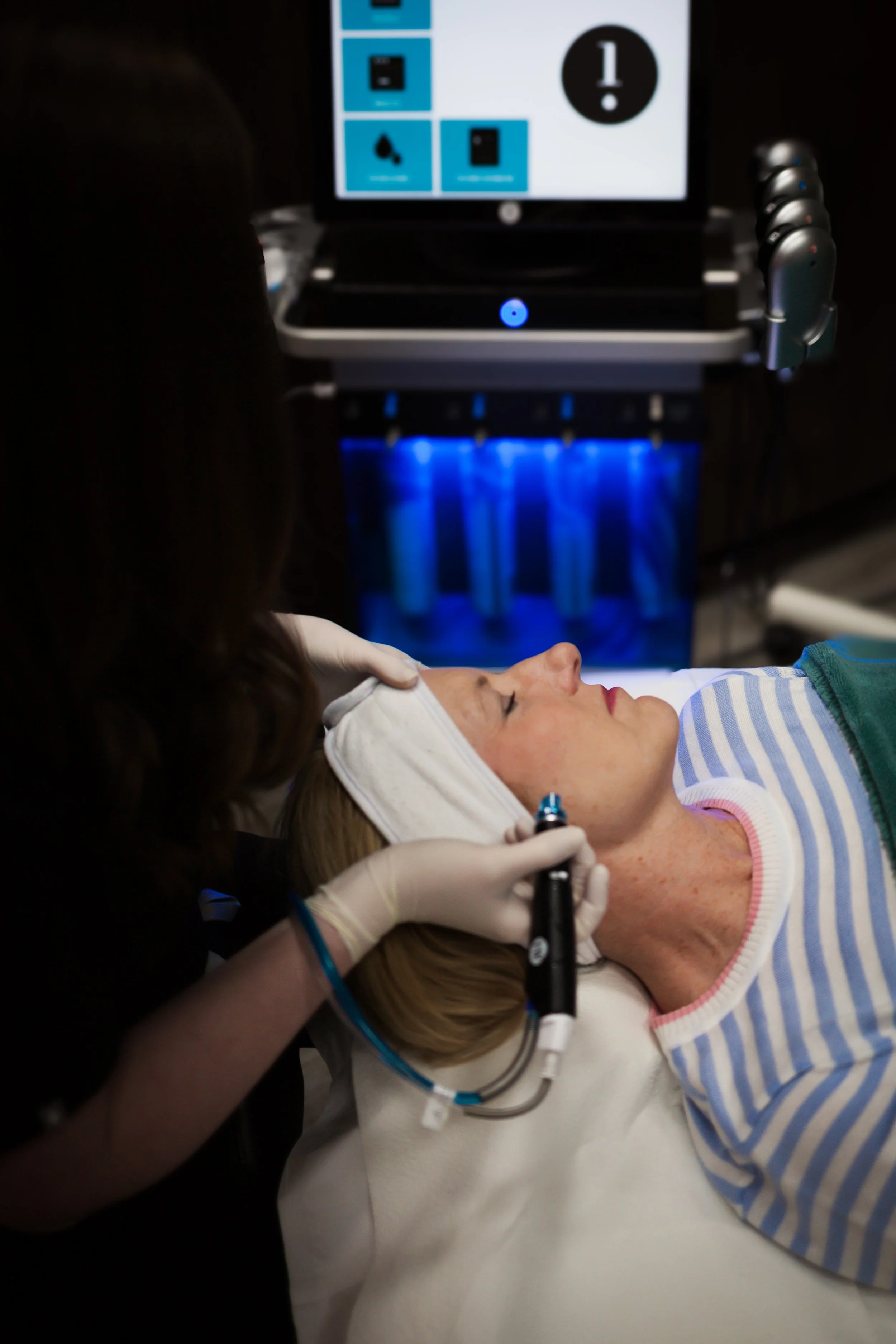 A woman lying on a hospital bed receiving a facial or medical treatment from a healthcare professional wearing gloves, with monitoring and medical equipment in the background.