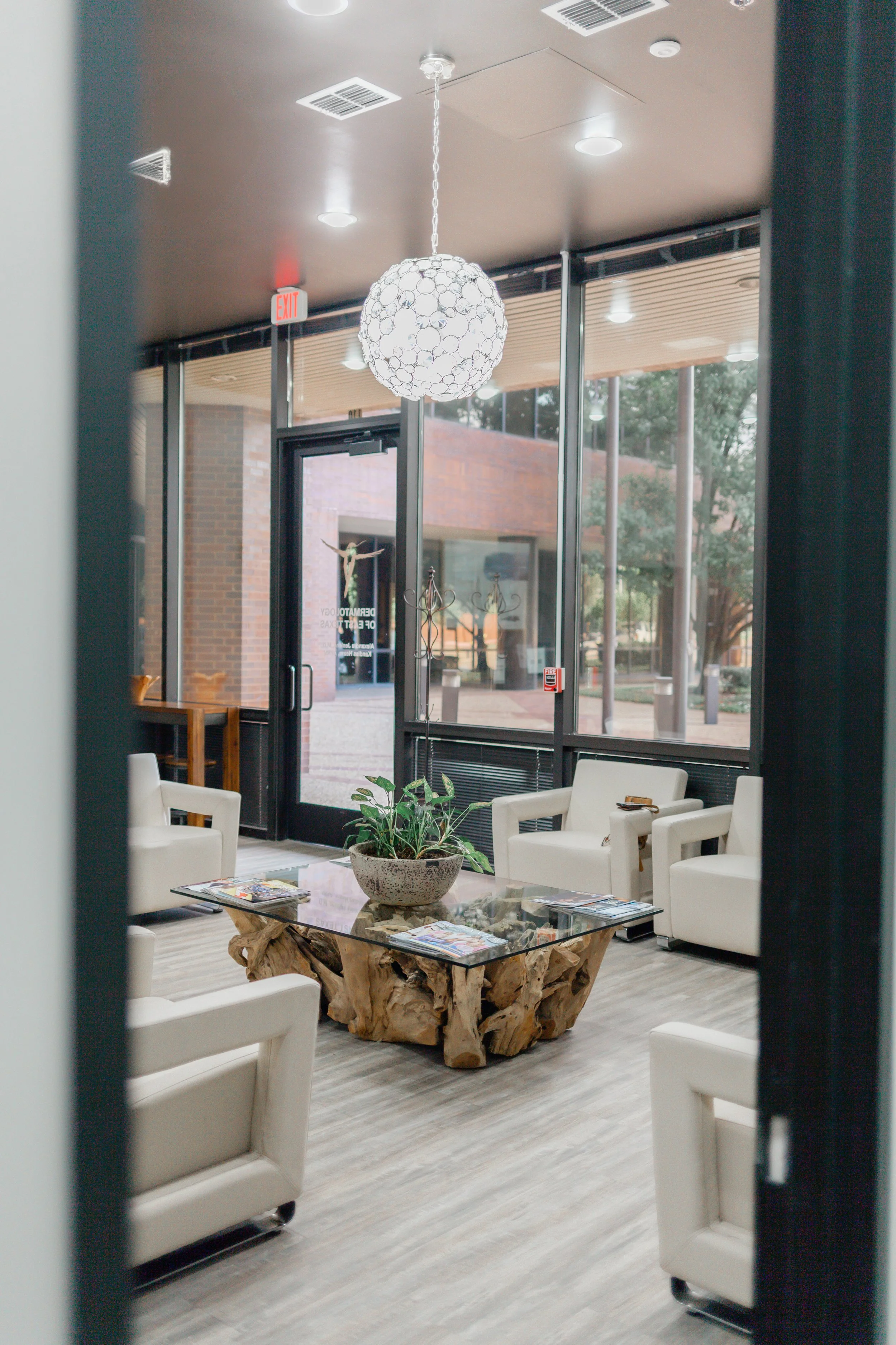 Dermatology of East Texas lobby with white armchairs, a wooden and glass table, potted plant, large glass windows, hanging spherical light fixture, and door with glass panels.