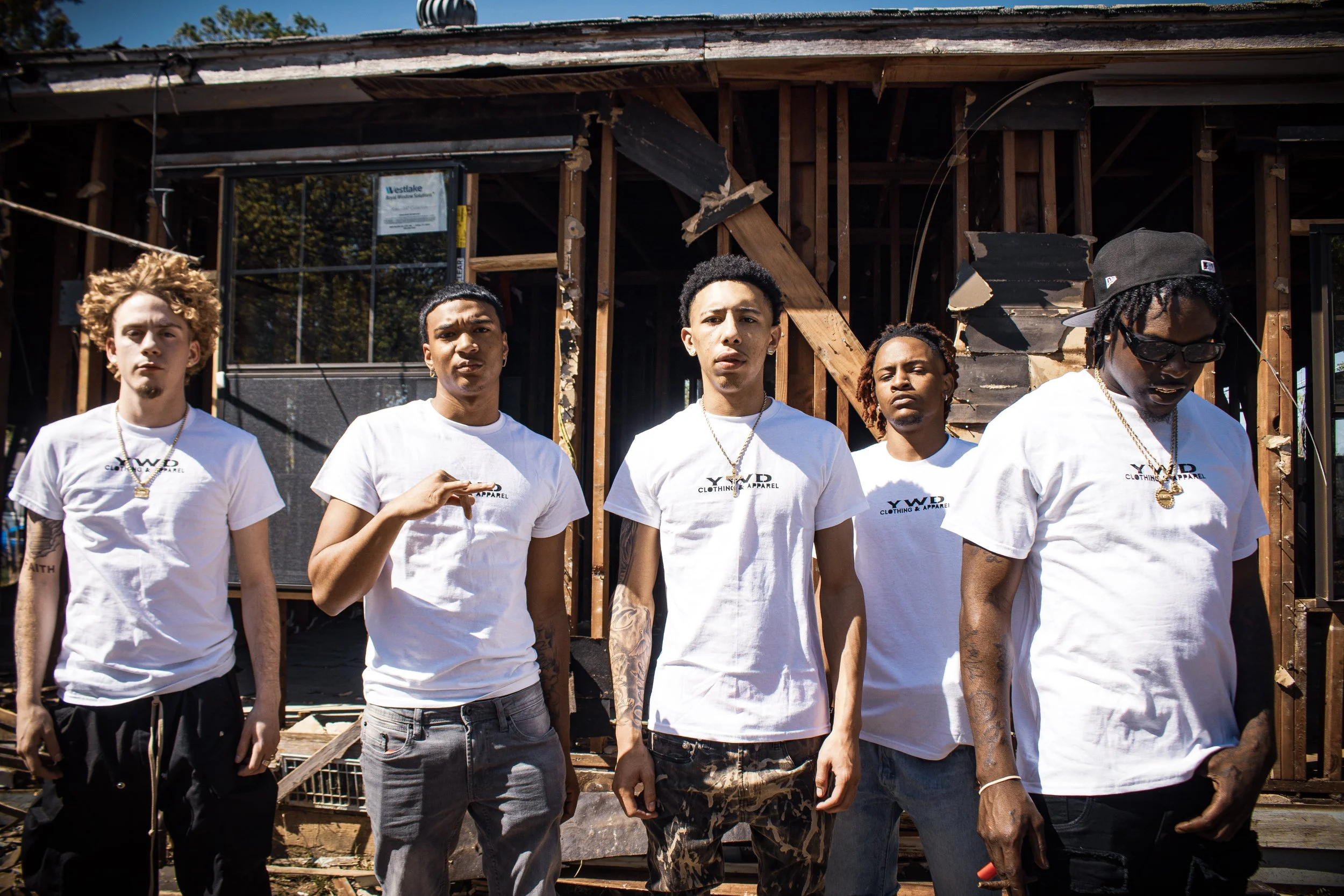 True Productions captures five young men (MYVSN) standing in front of a partially constructed building during daytime. They are wearing white T-shirts with a logo, some have tattoos, jewelry, and accessories, and are looking serious or pensive.