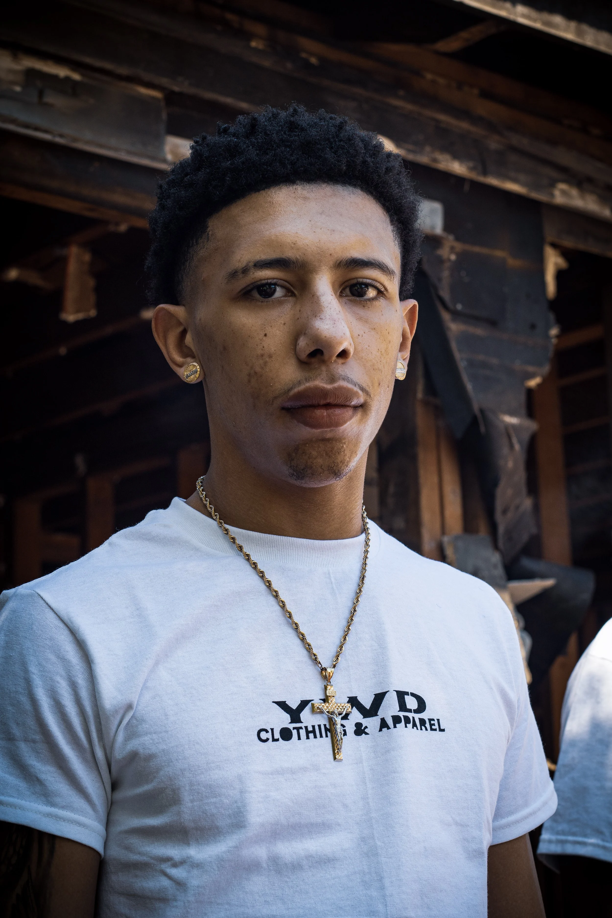 Portrait of a young man with short curly hair, wearing a white T-shirt with black text, gold earrings, and a gold chain with a pendant. Background shows a wooden structure.
