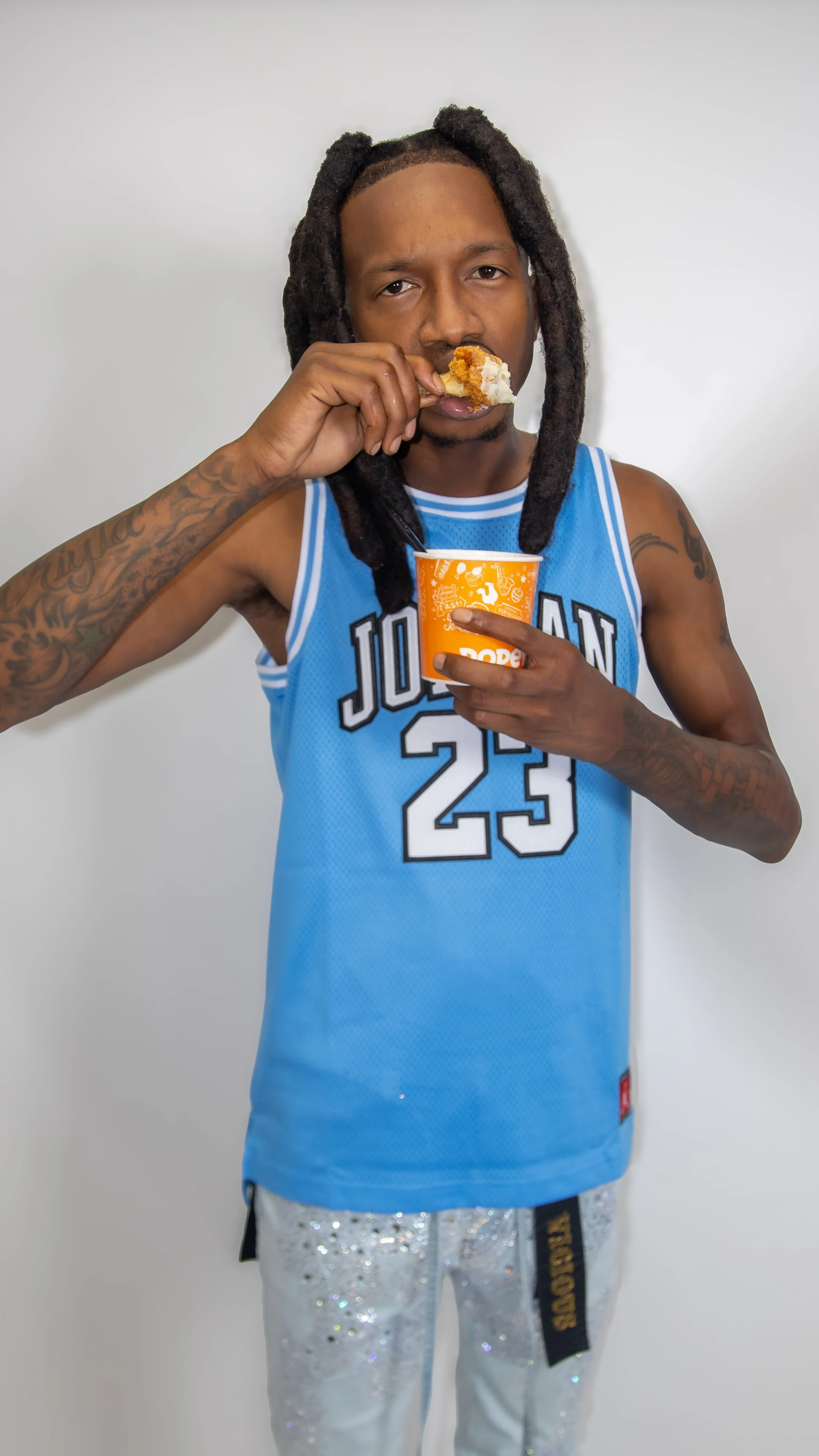 A man with dreadlocks wearing a blue basketball jersey and sequined pants, eating food from a cup and holding a piece of fried chicken.