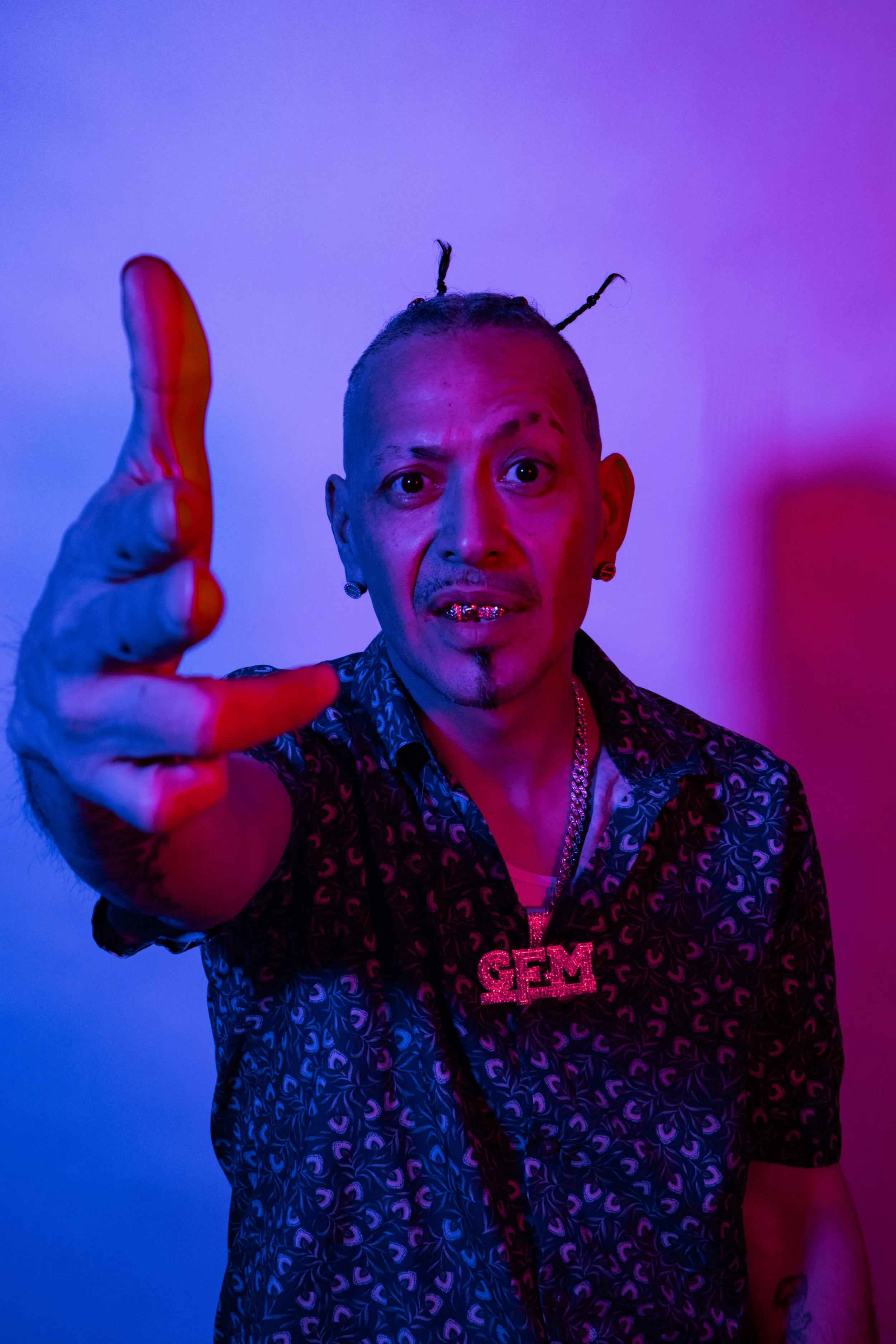 A man with styled hair, gold grill on teeth, wearing a patterned shirt, and a glittery 'GEM' necklace, reaching toward the camera with a colorful pink and blue lighting background.