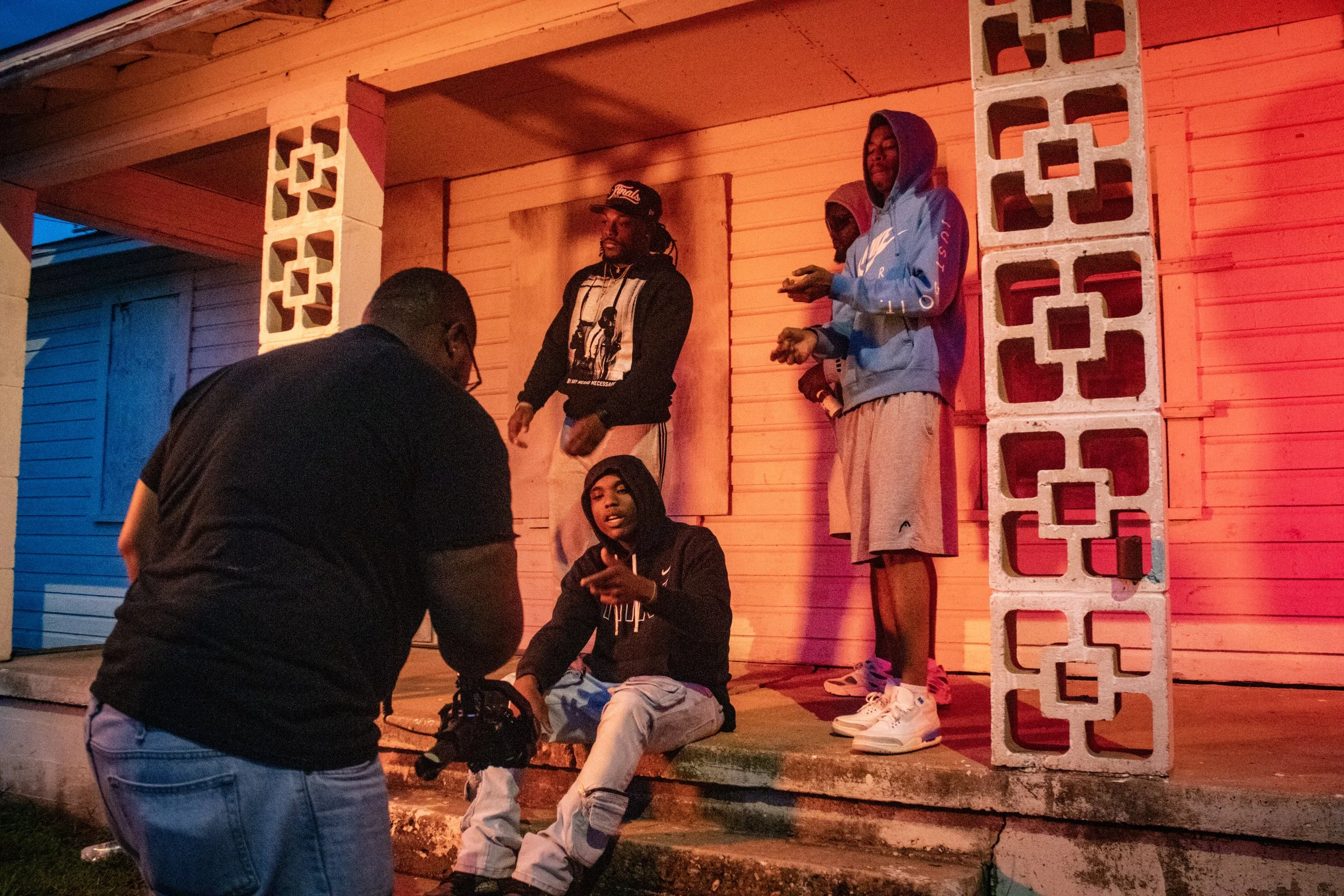 A group of young men are gathered on a porch at night, with some sitting and others standing, illuminated by warm lighting. One man is sitting while another appears to be filming or photographing him. The porch has wooden siding and concrete blocks as part of the structure.