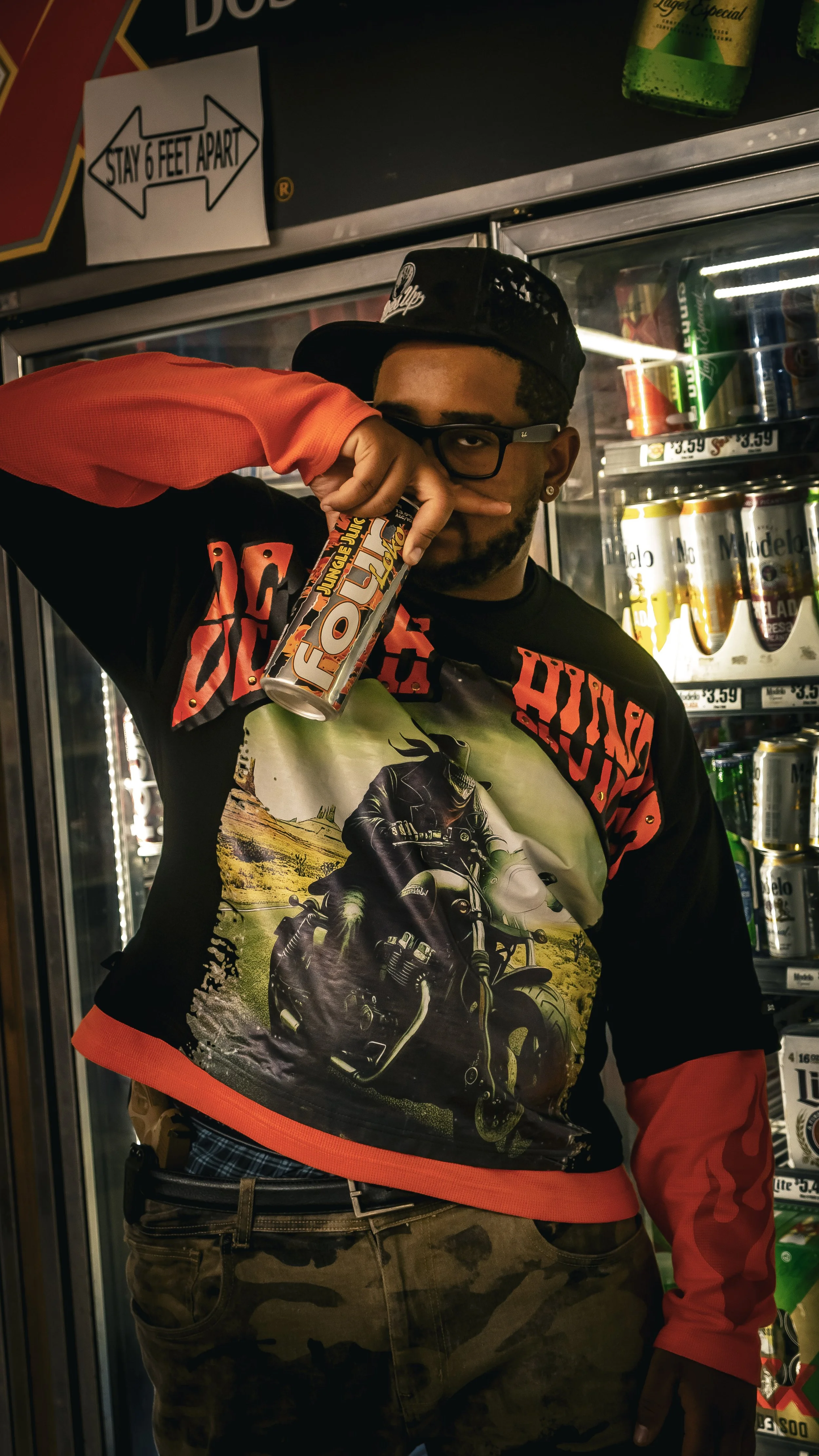 A man wearing glasses, a backwards black cap, and a graphic motorcycle-themed shirt standing in front of a convenience store refrigerator. He is holding a can of FOLT energy drink near his face.