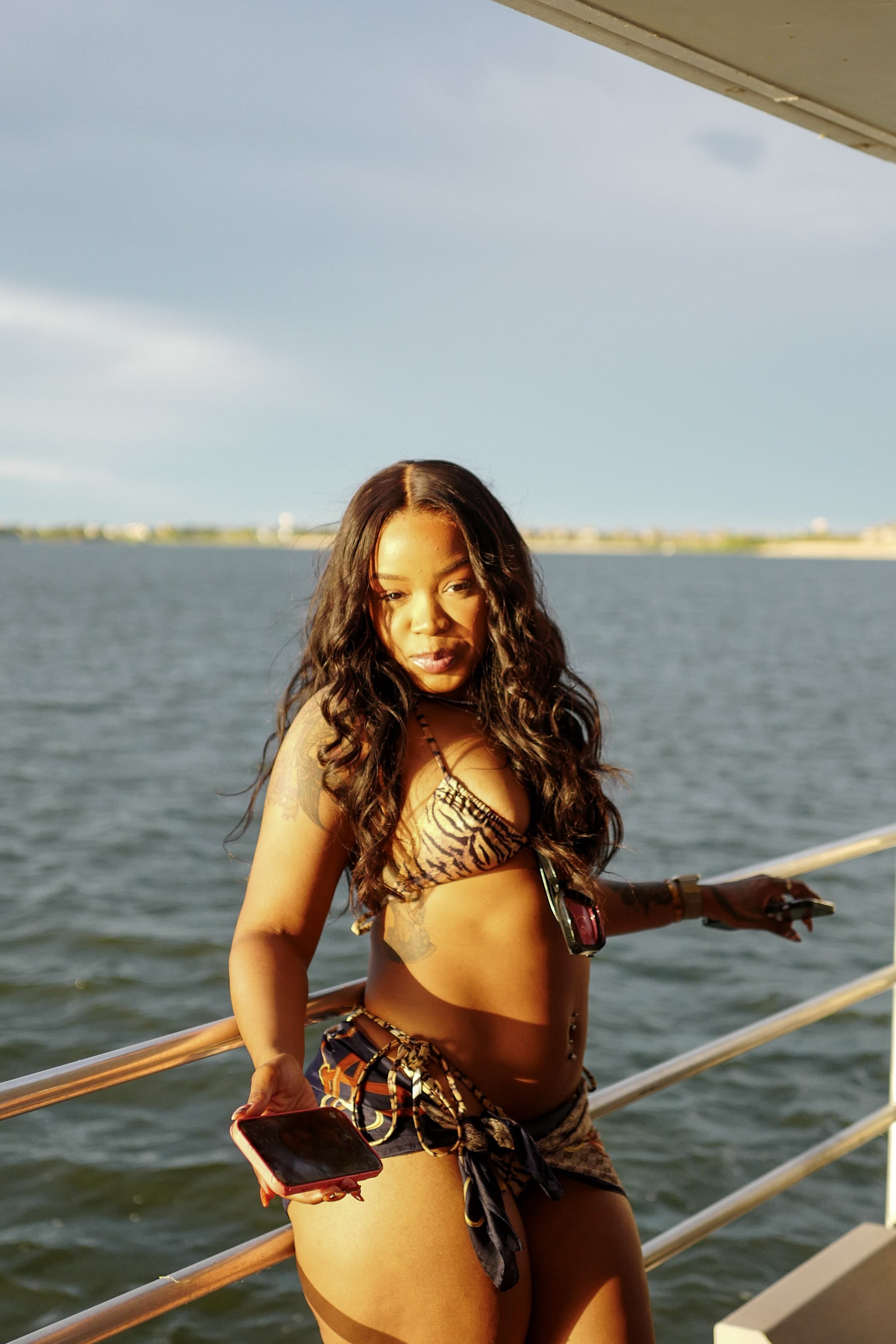 A woman with long curly hair standing on a boat deck by the water, holding a smartphone in one hand, wearing a tiger-print bikini top and matching shorts, with tattoos on her arm and a small glasses hanging from her bikini top.