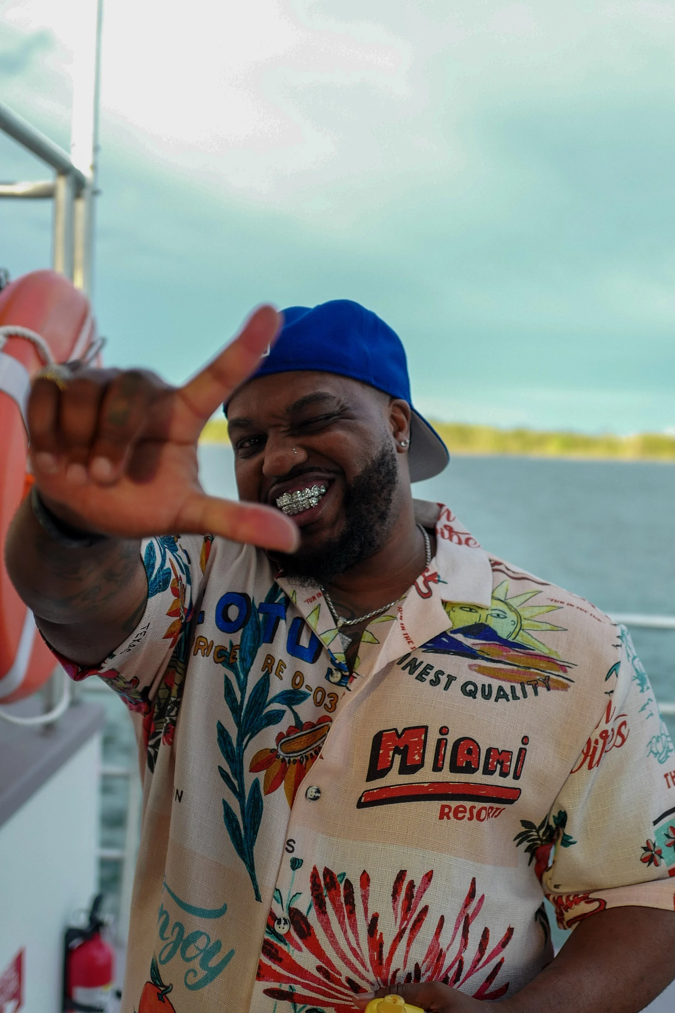 A man on a boat, wearing a colorful tropical shirt and a blue baseball cap, making a hand gesture with his left hand while winking and smiling with grills on his teeth. The background shows water and a cloudy sky.