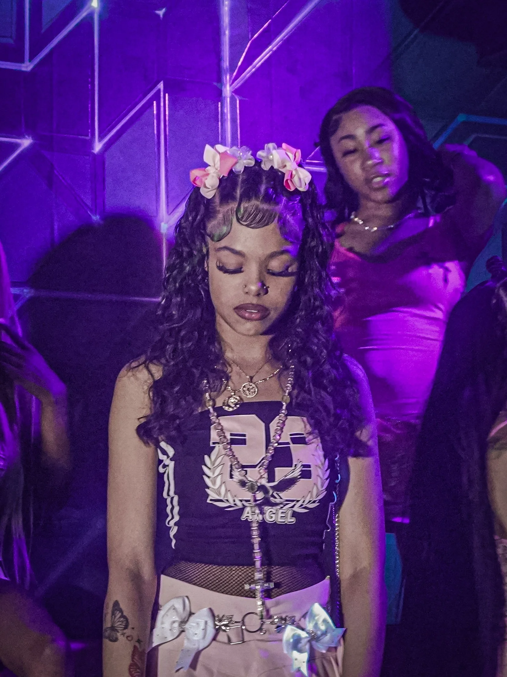 Young woman with curly hair and floral headband looking down, wearing a black and white tank top with 'Angel' text, layered necklaces, and pastel pink pants at a neon-lit party.