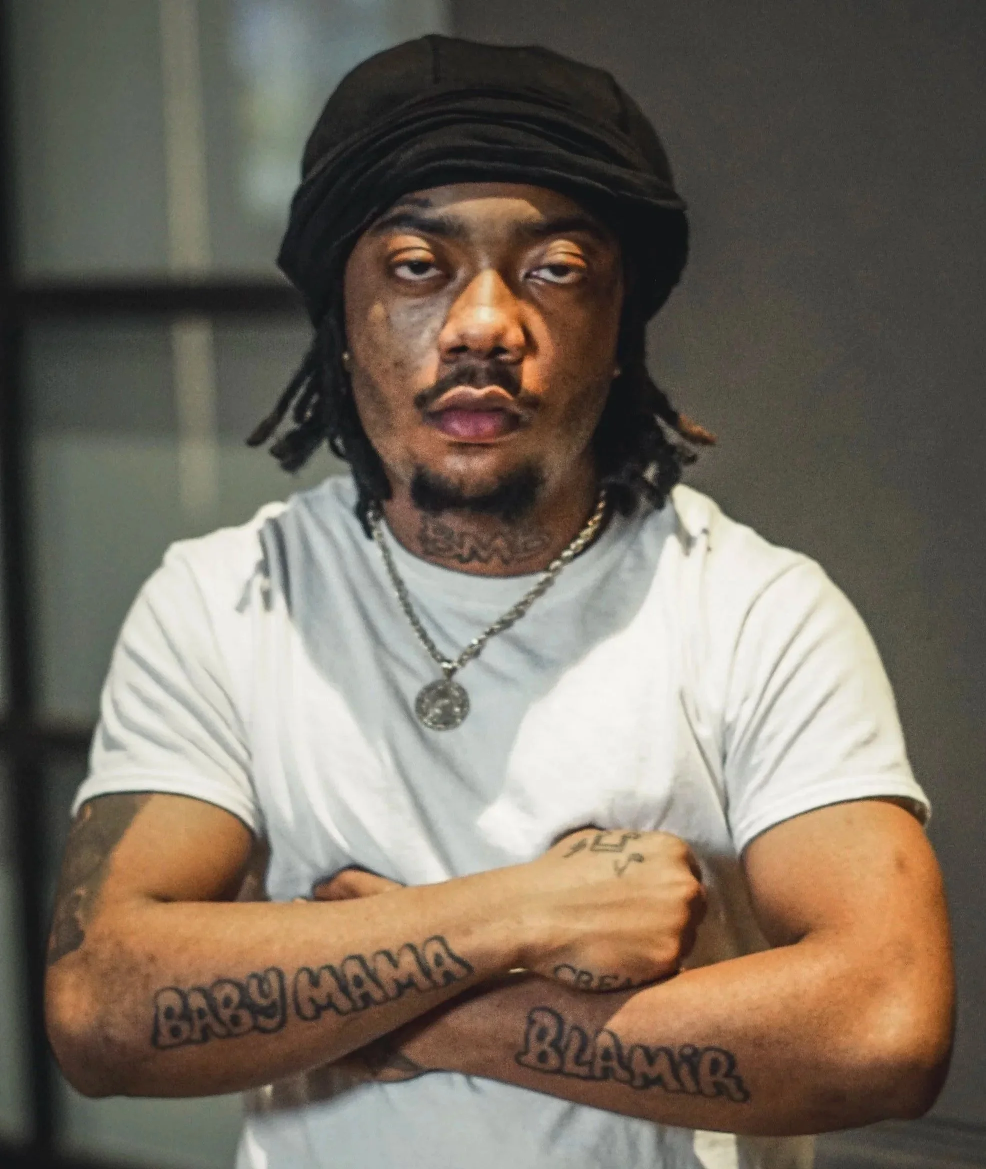 Young man with dreadlocks wearing a black headwrap, white t-shirt, and necklaces, crossing his arms, showing tattoos on both arms that say 'Baby Mama' and 'Blaque', with a serious expression.