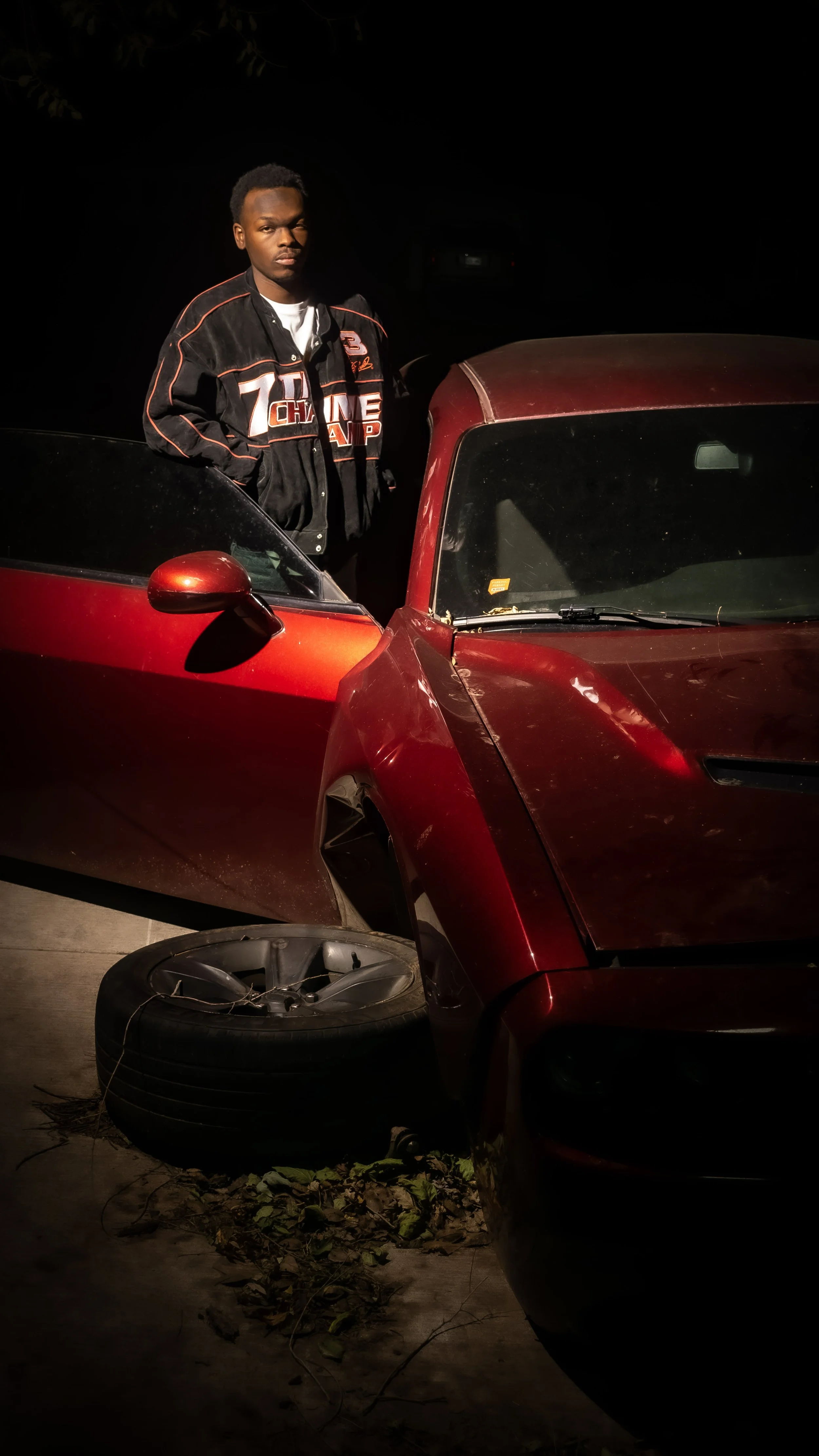A young man standing next to a red sports car at night. The car appears to be parked with a flat tire, and a spare tire is lying on the ground nearby. The young man is wearing a black jacket with red and white graphics.