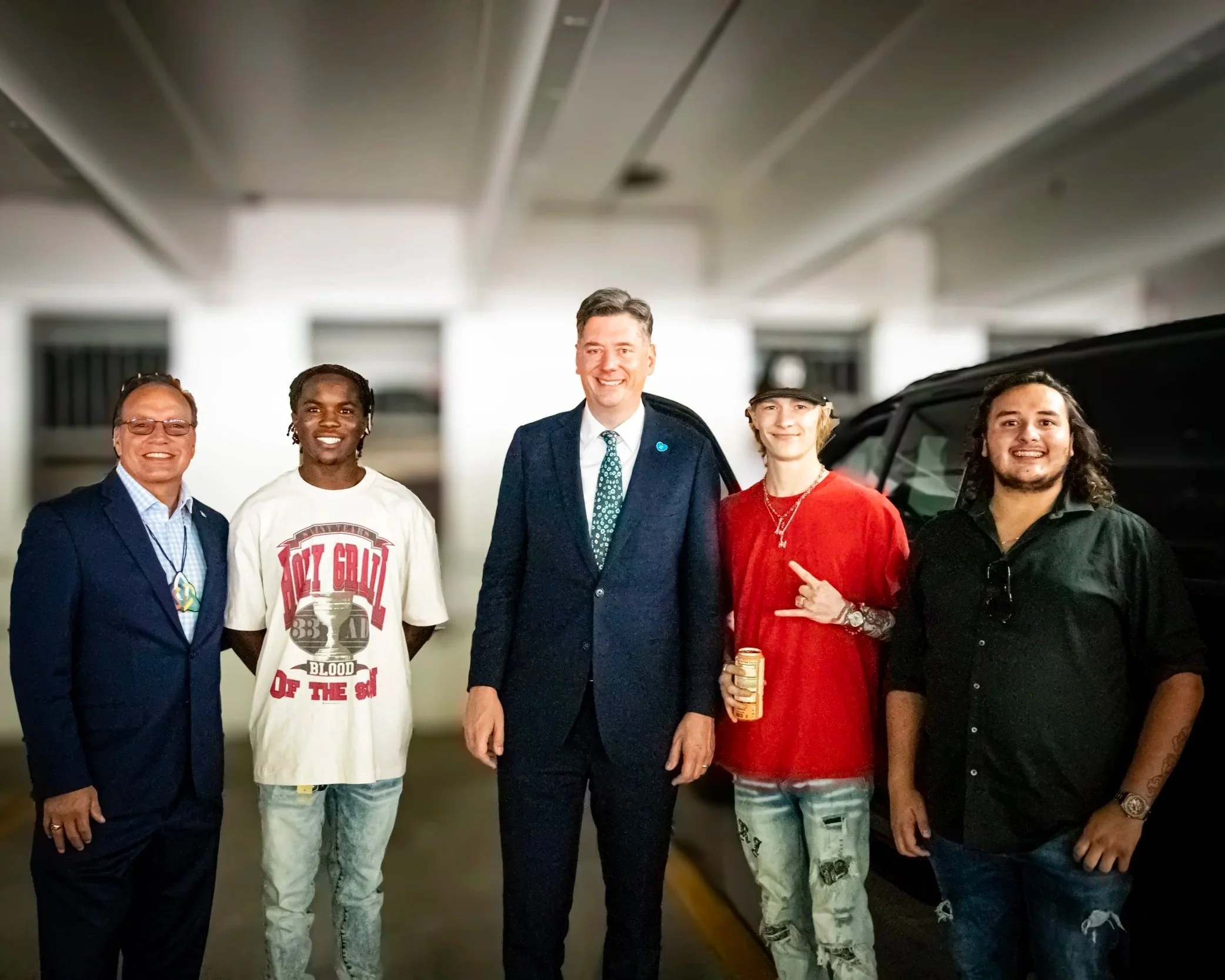 True Productions captures a group of four men with OKC mayor David Holt standing in a parking garage, smiling at the camera. They are dressed in casual and business attire, with one wearing a graphic t-shirt, and three wearing suits or shirts.