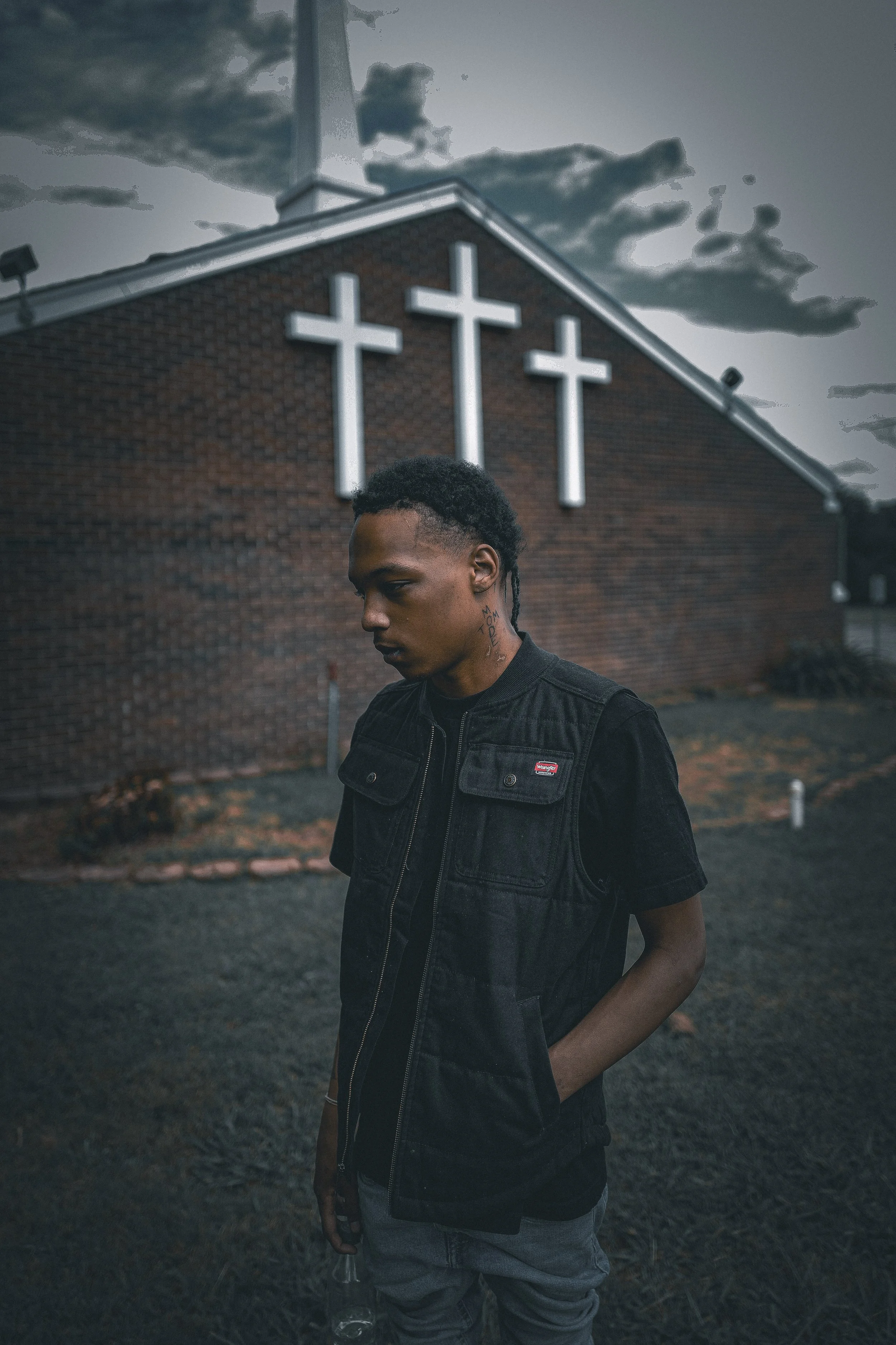 A young man in a black denim vest standing outside of a church with three large white crosses on the brick wall behind him during dusk.