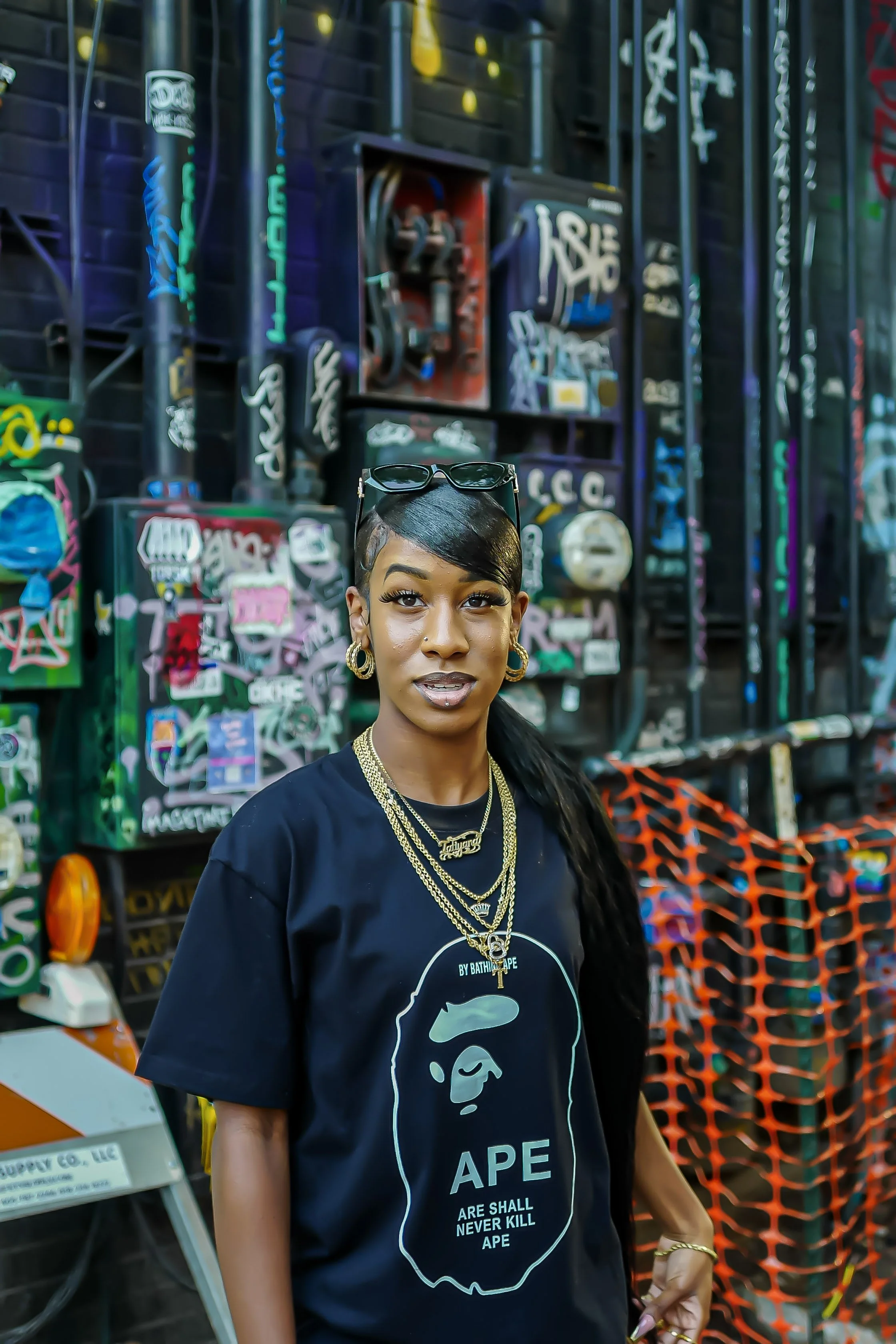 True Productions captures a woman wearing a black BAPE t-shirt with a graphic and text, standing in front of a graffiti-covered wall filled with various wires, boxes, and street art.