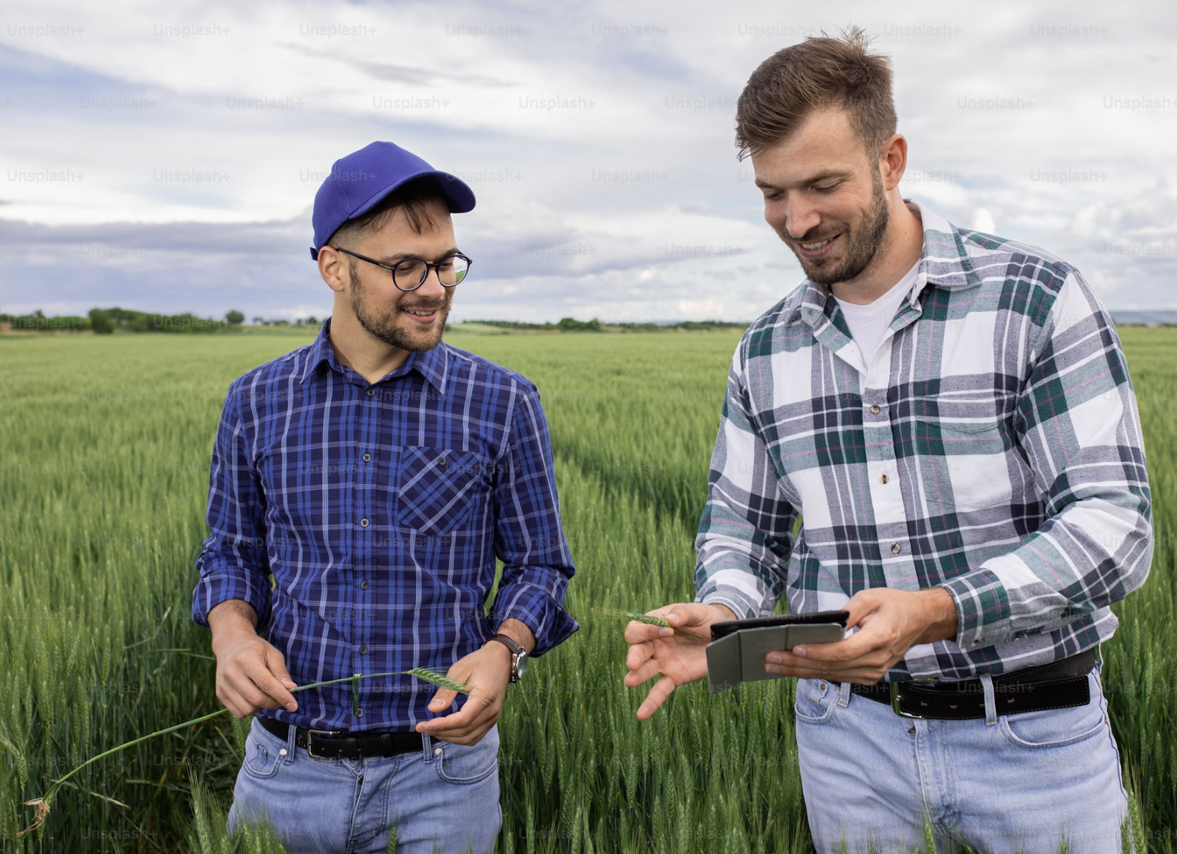 Deux hommes dans un champ verdoyant, regardant un appareil électronique, souriant, sous un ciel nuageux.