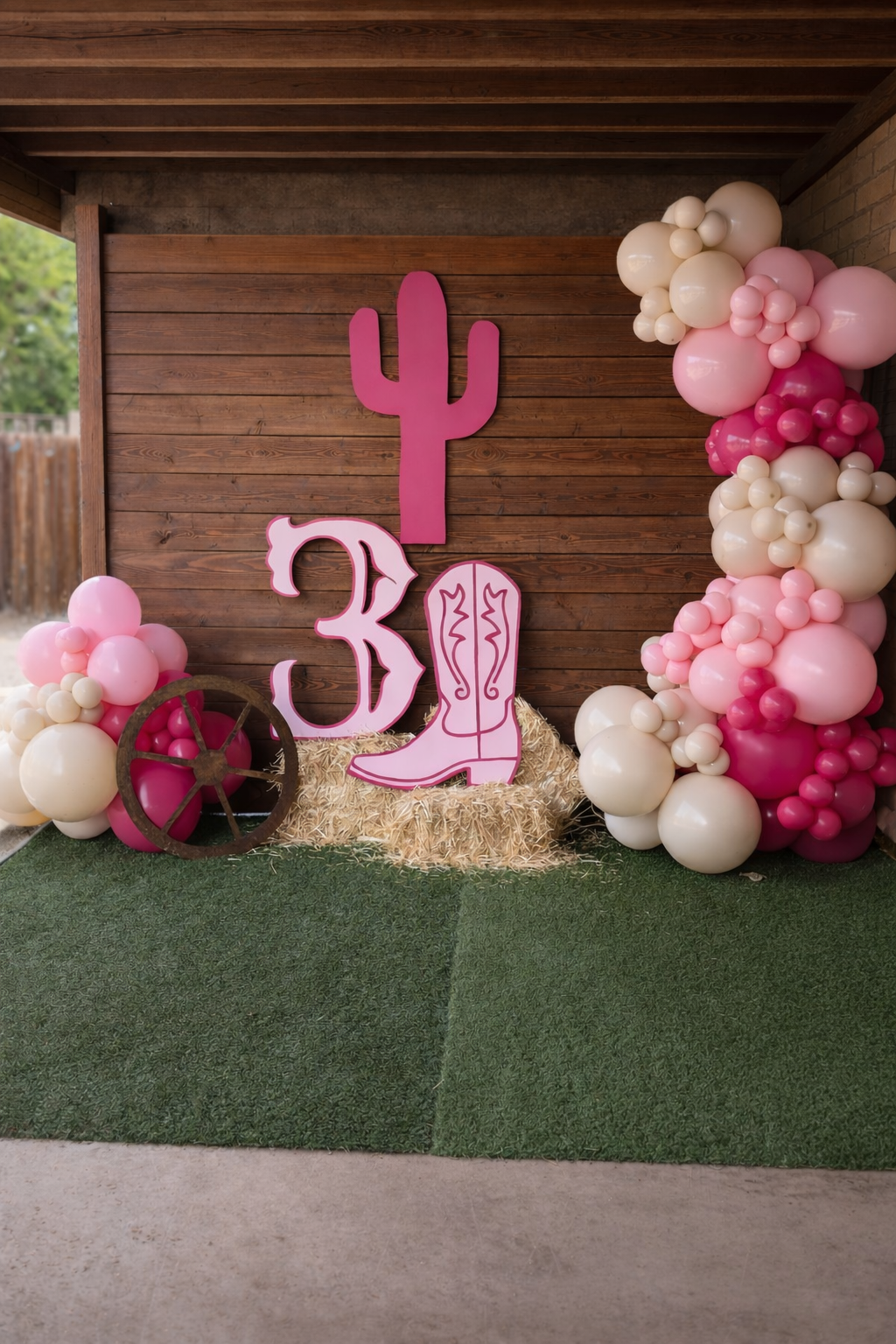 Pink cowboy-themed birthday party decoration with balloons, a large pink cactus cutout, a pink cowboy boot cutout, and a wagon wheel, set against a wooden backdrop.
