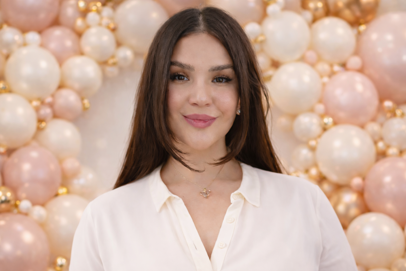 A woman with long dark hair, wearing a white blouse and a heart-shaped necklace, standing in front of a backdrop decorated with pink, white, and gold balloons.