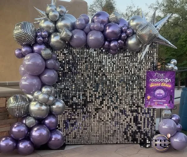 Decorative backdrop with purple, silver, and checkered balloons forming an arch, a sparkling silvery curtain in the background, and a sign for the TMC Radiothon Silent Auction.