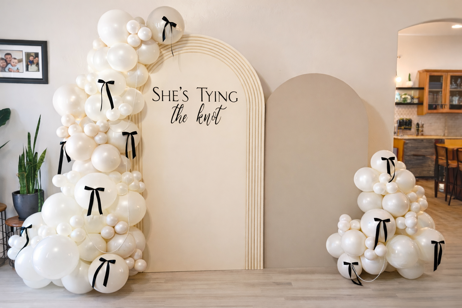 Decorative wedding backdrop with white balloons adorned with black bows, arranged in clusters on the left and right sides, with the phrase "She's Tying the Knot" written on an arch-shaped panel in the center, inside a home with a kitchen visible in the background.