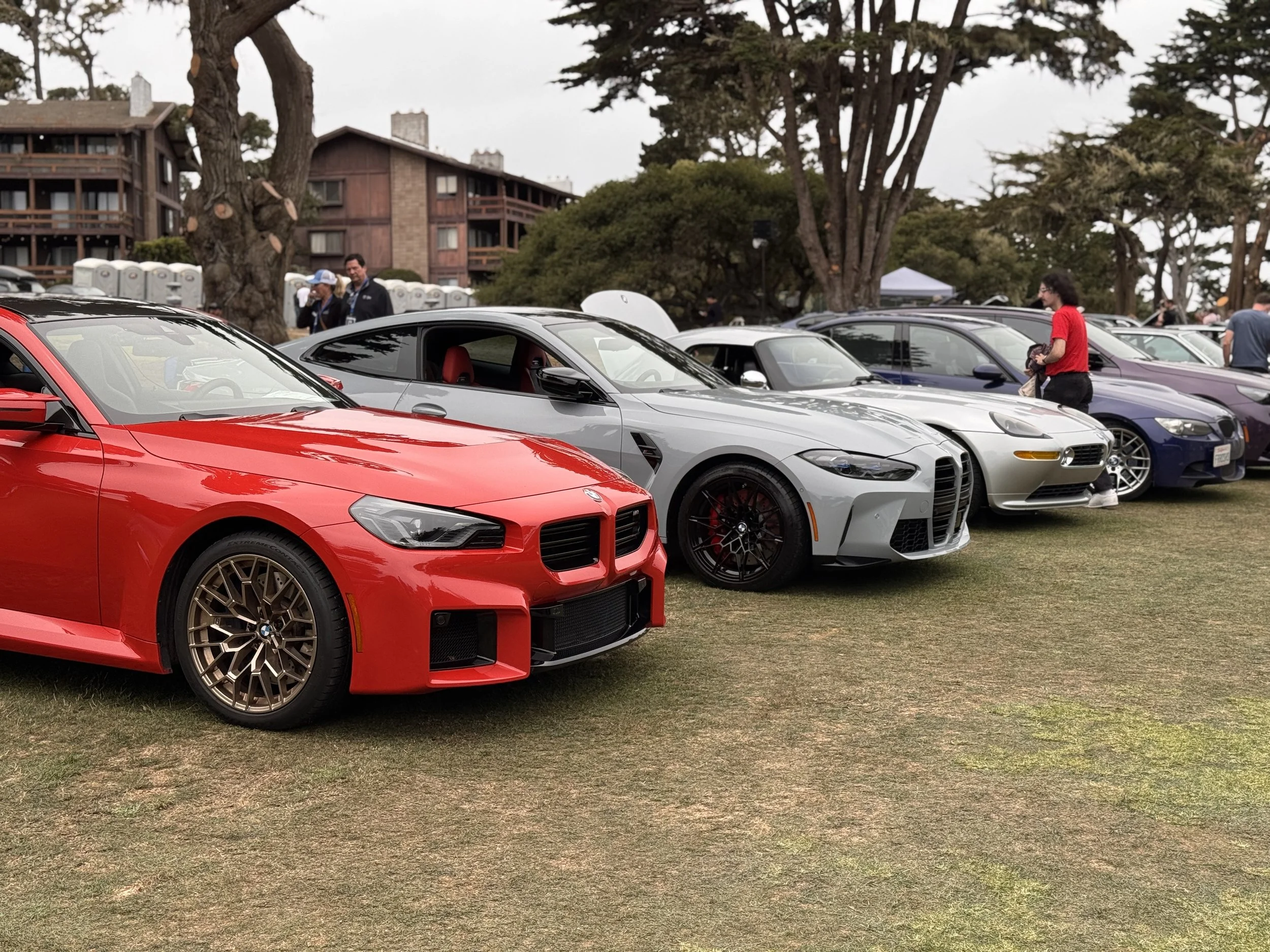 Lineup of various sports cars on a grassy field during an outdoor car show, with attendees inspecting and admiring the vehicles.