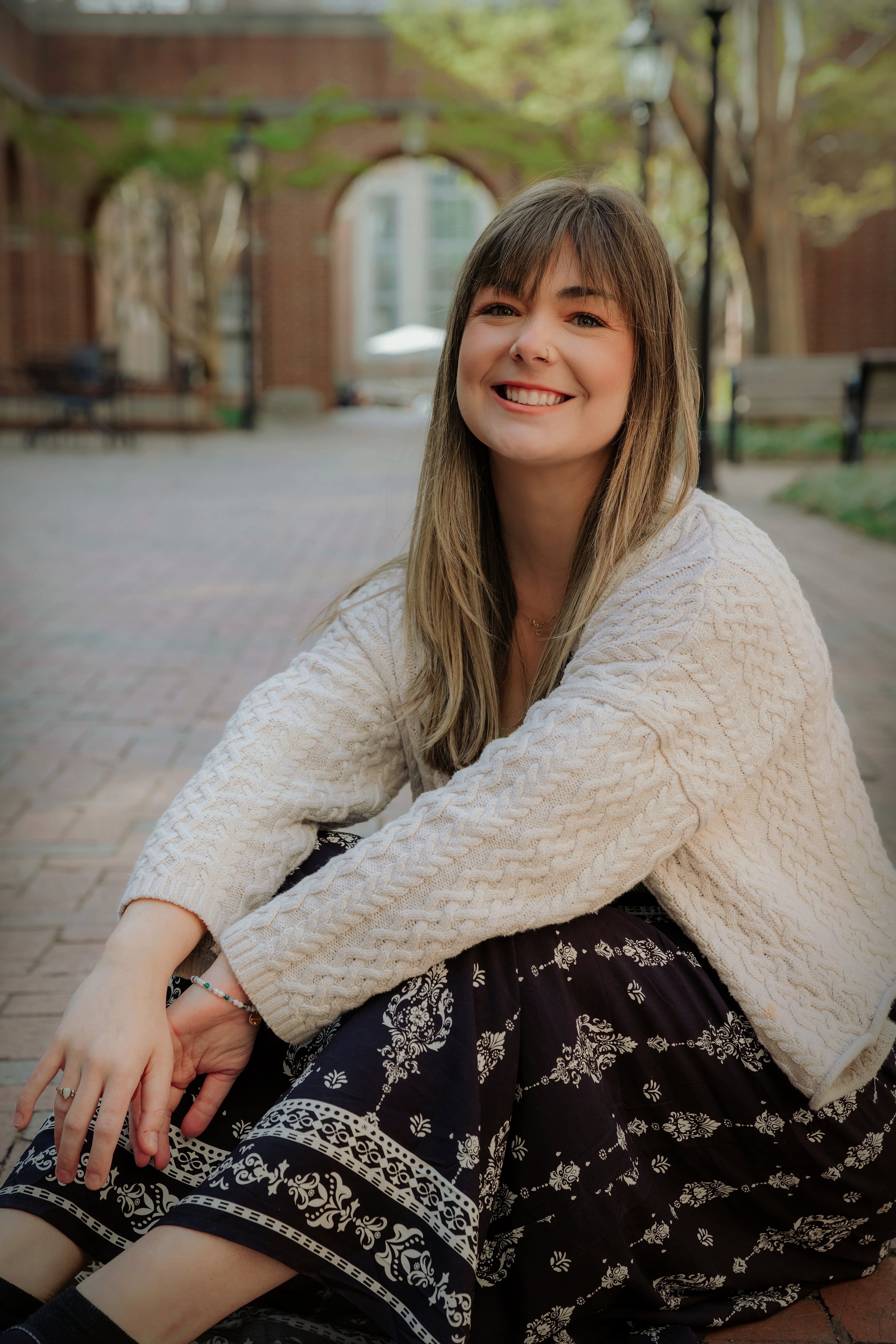 Young female presenting therapist sitting outdoors on a brick pathway, smiling, wearing a cream cable-knit sweater and patterned black skirt, with trees, benches, and brick arches in the background.