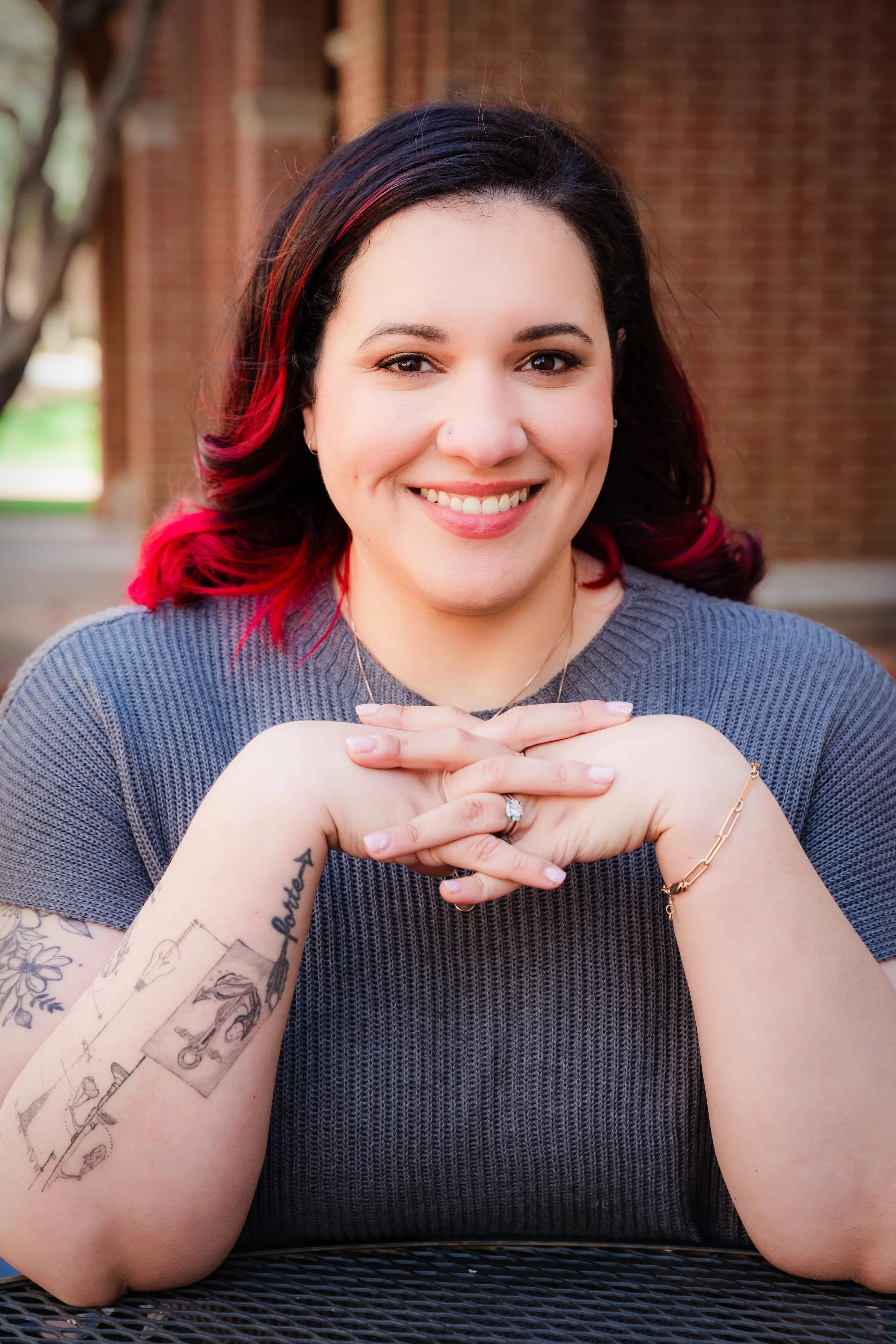 A woman smiling outdoors with black and red hair, wearing a gray shirt, resting her chin on her clasped hands, with tattoos on her left arm, a necklace, and a ring on her finger, in front of a brick building.