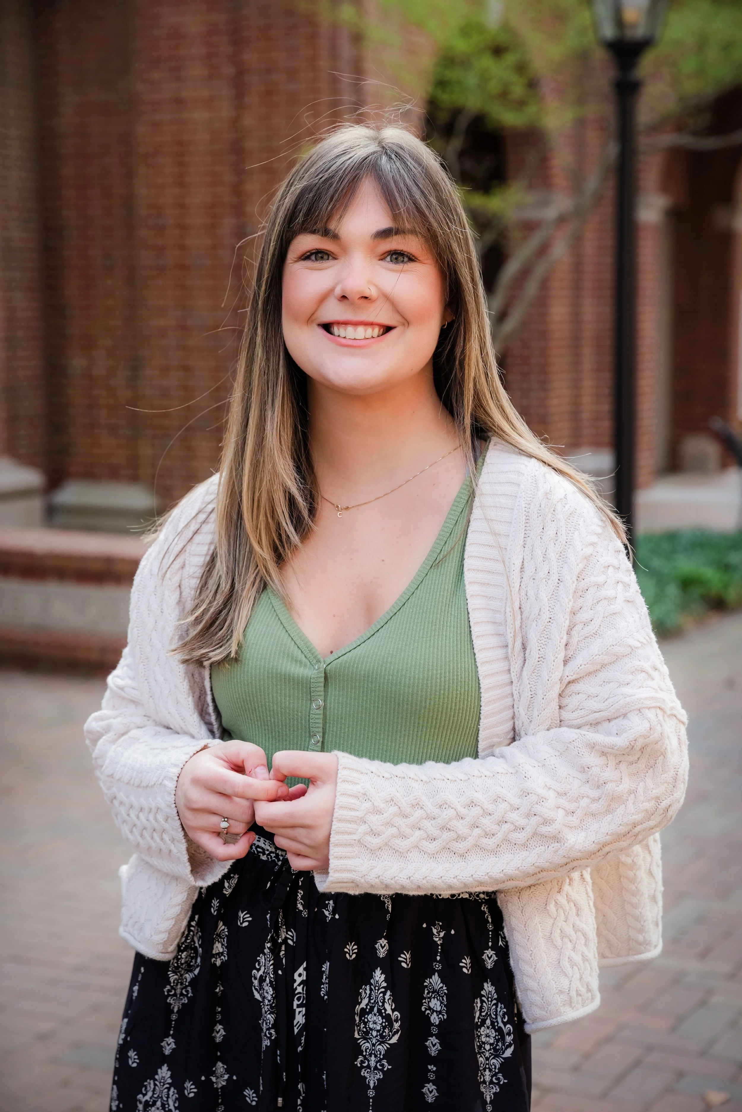 Smiling woman standing outdoors in front of a brick wall, wearing a green top, beige cardigan, and black skirt, with trees and street lamps in the background.