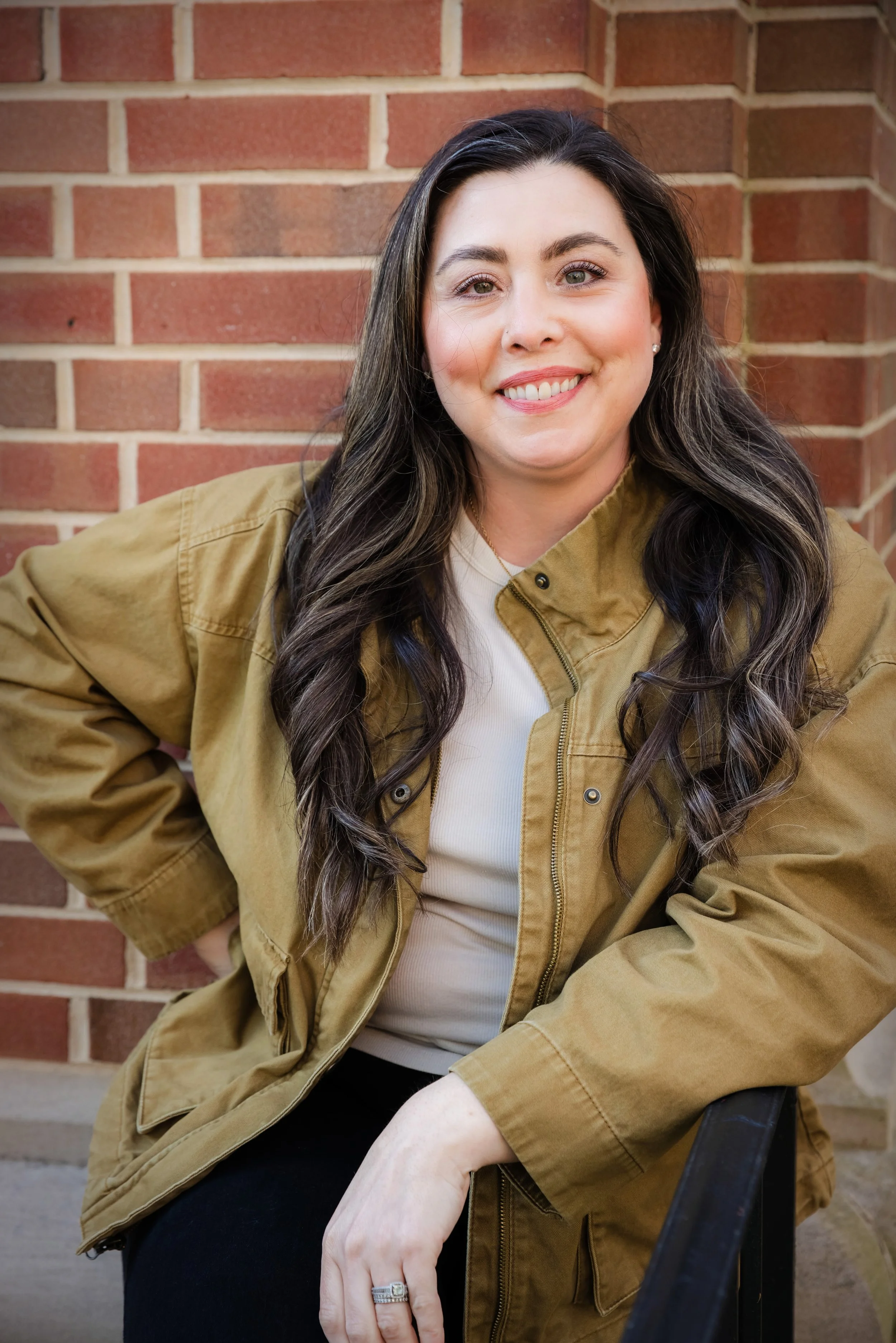 A woman with long, wavy dark hair smiling, wearing a tan jacket over a white top, sitting against a brick wall.