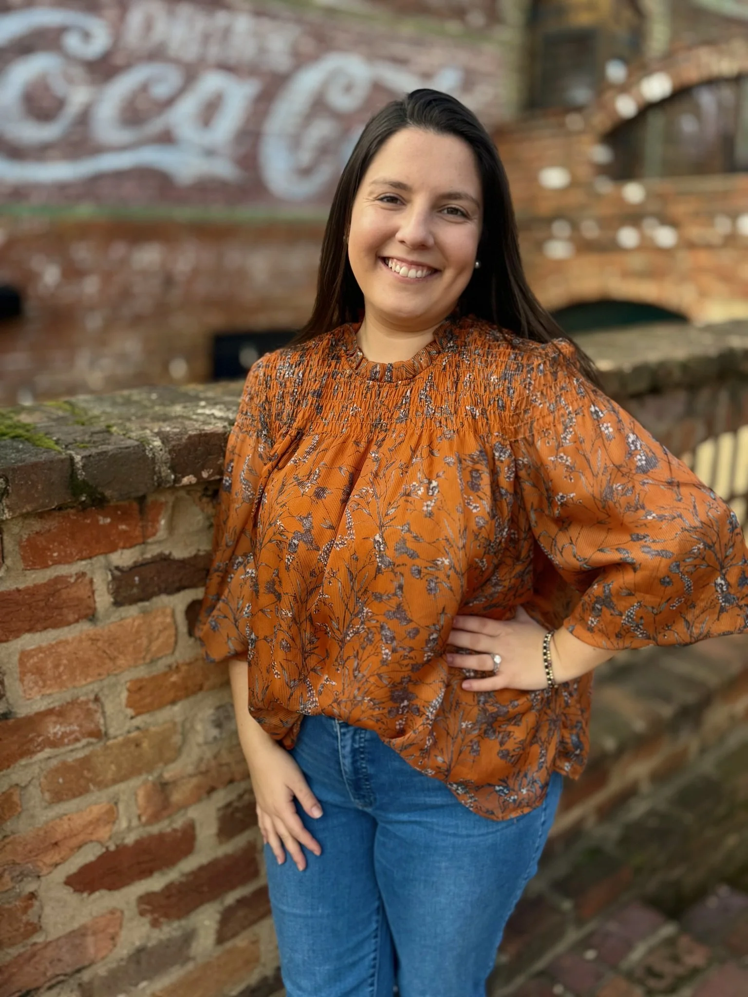 A woman with long dark hair smiling, wearing an orange blouse with floral patterns and blue jeans, standing outdoors in front of a brick wall with a painted sign in the background.