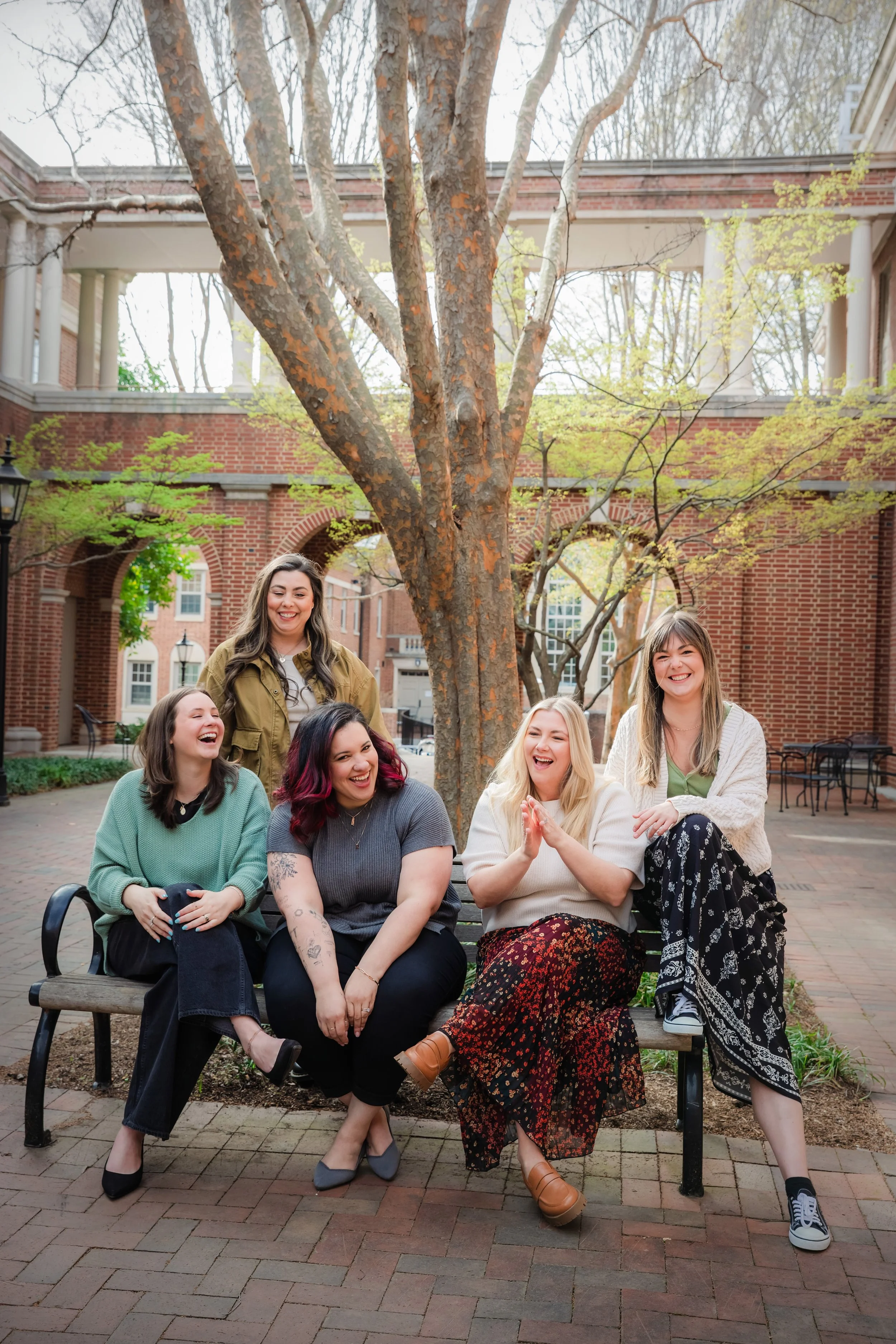 A group of six diverse women sitting and standing on a park bench in an outdoor brick courtyard with trees, brick buildings, and a stone walkway. They are smiling and laughing, enjoying each other's company.