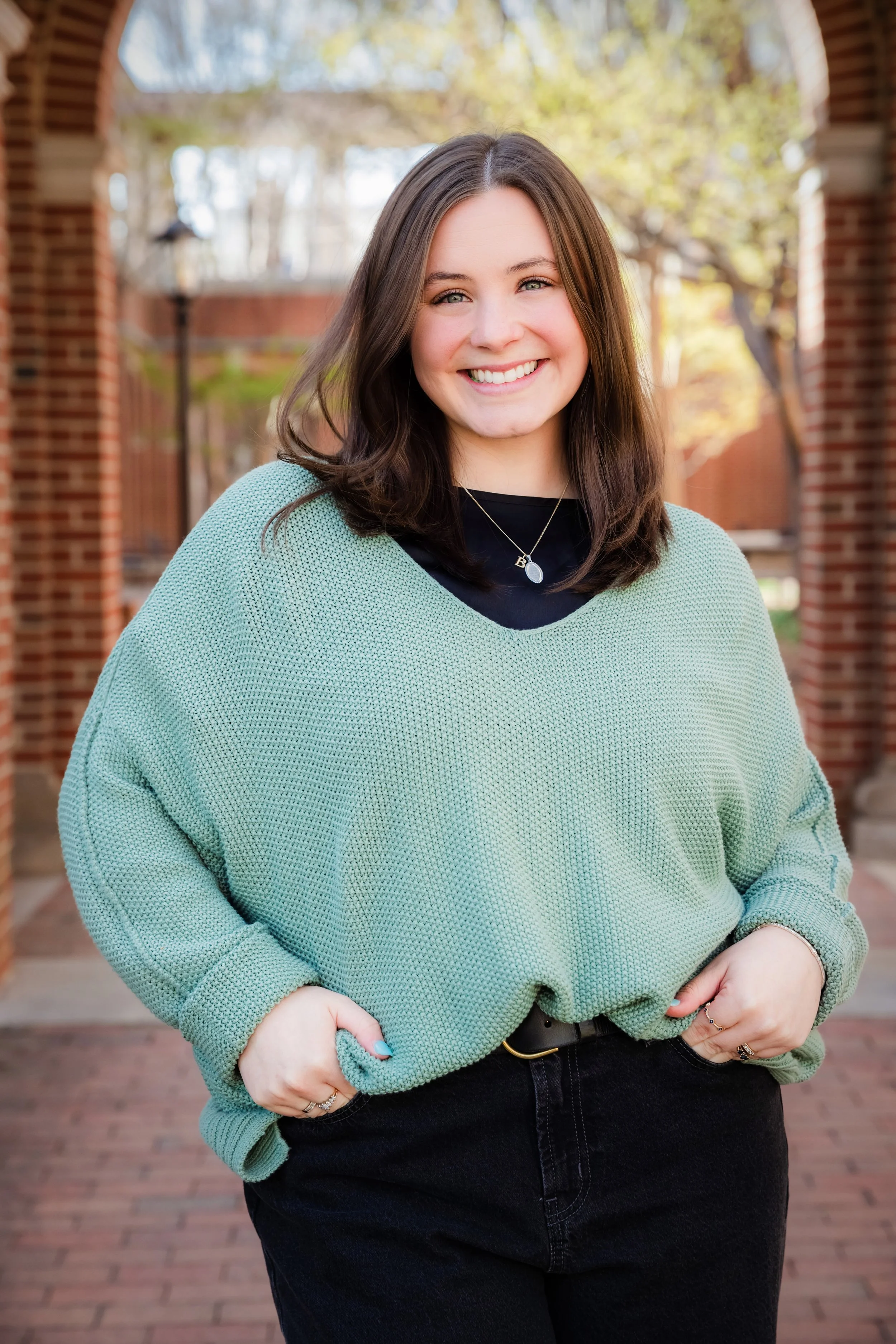 Smiling woman with brown hair wearing a mint green sweater, black shirt, and a necklace, standing outdoors with trees and brick structures in the background.