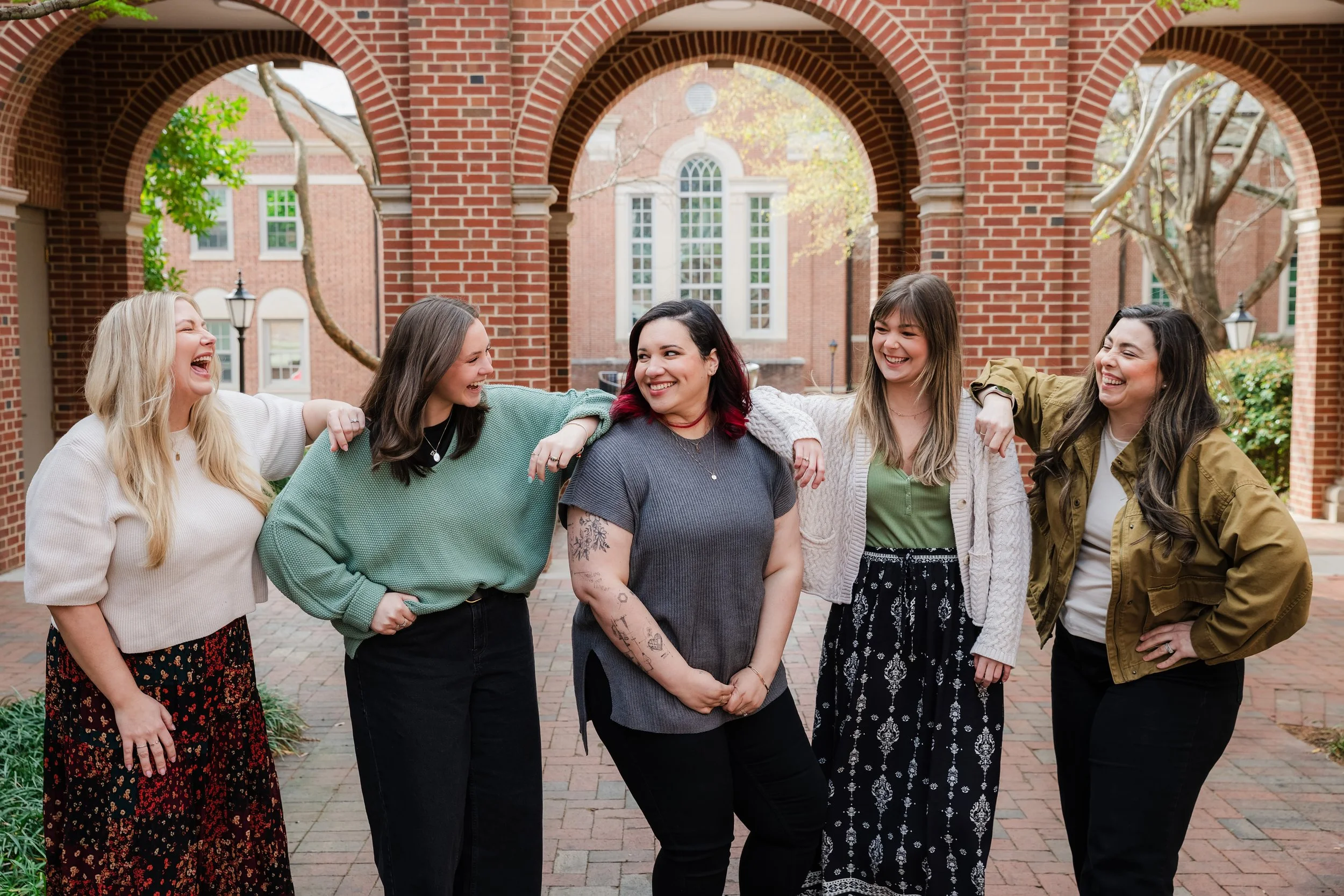 A group of six female presenting therapists standing together outdoors, smiling, with brick arches and trees in the background.