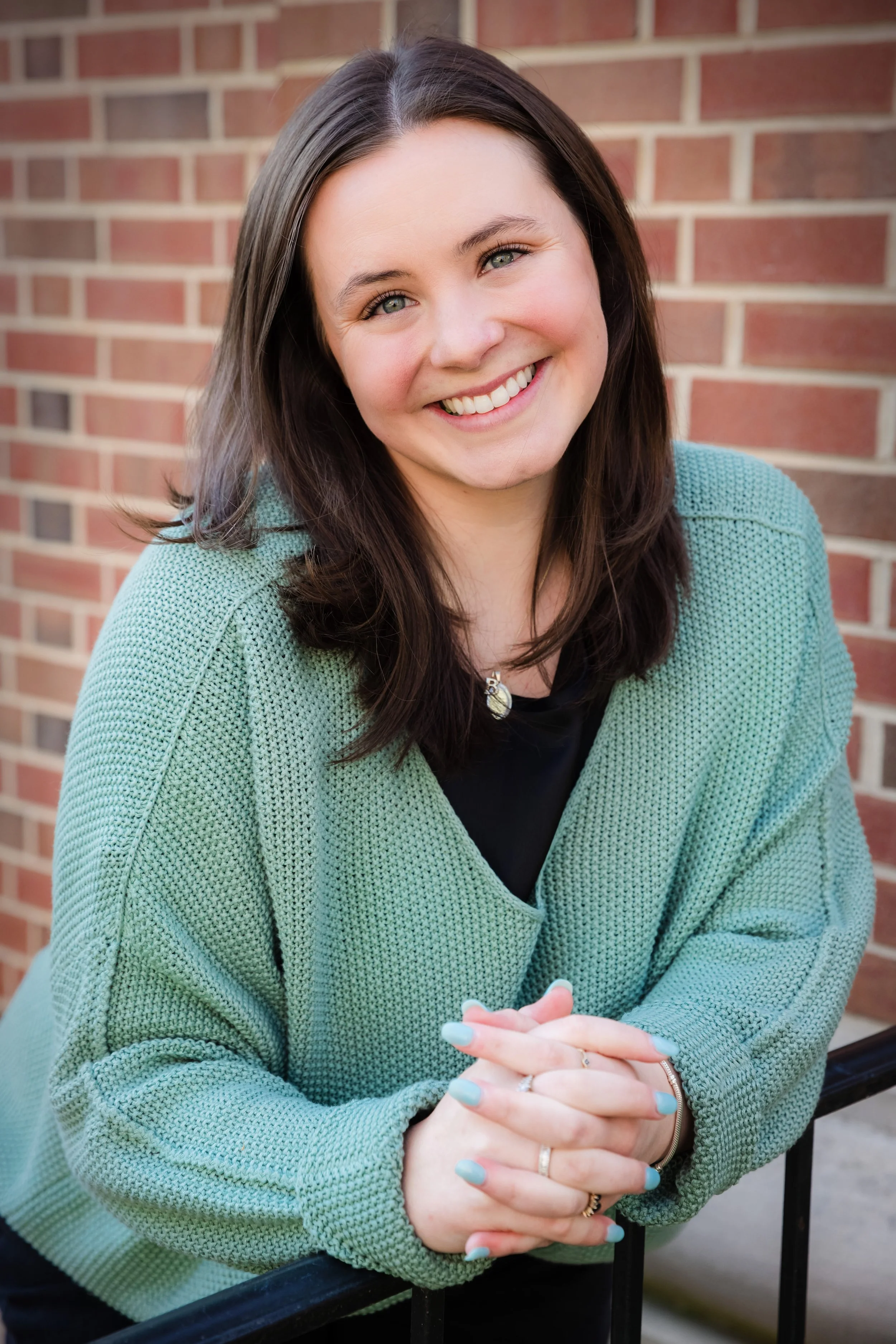 A young woman with shoulder-length dark brown hair and light skin, smiling, wearing a green sweater with a black shirt underneath, and posed outdoors in front of a brick wall.