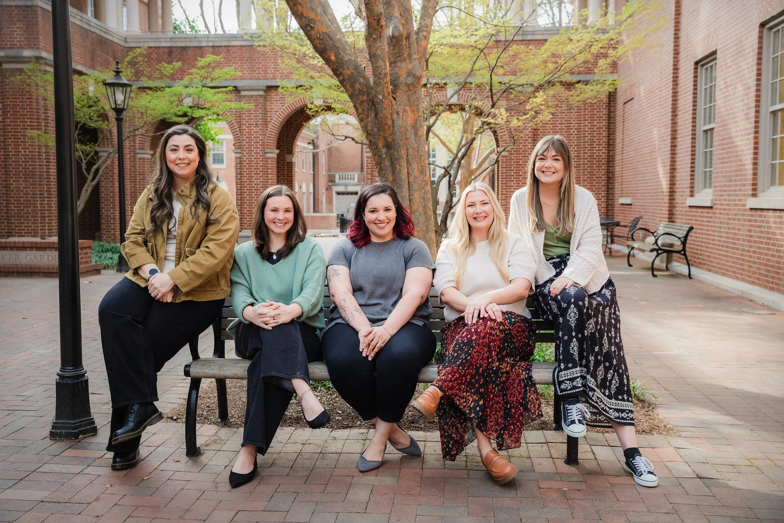 Five women sitting on a bench in front of a brick building with trees and benches, smiling at the camera.