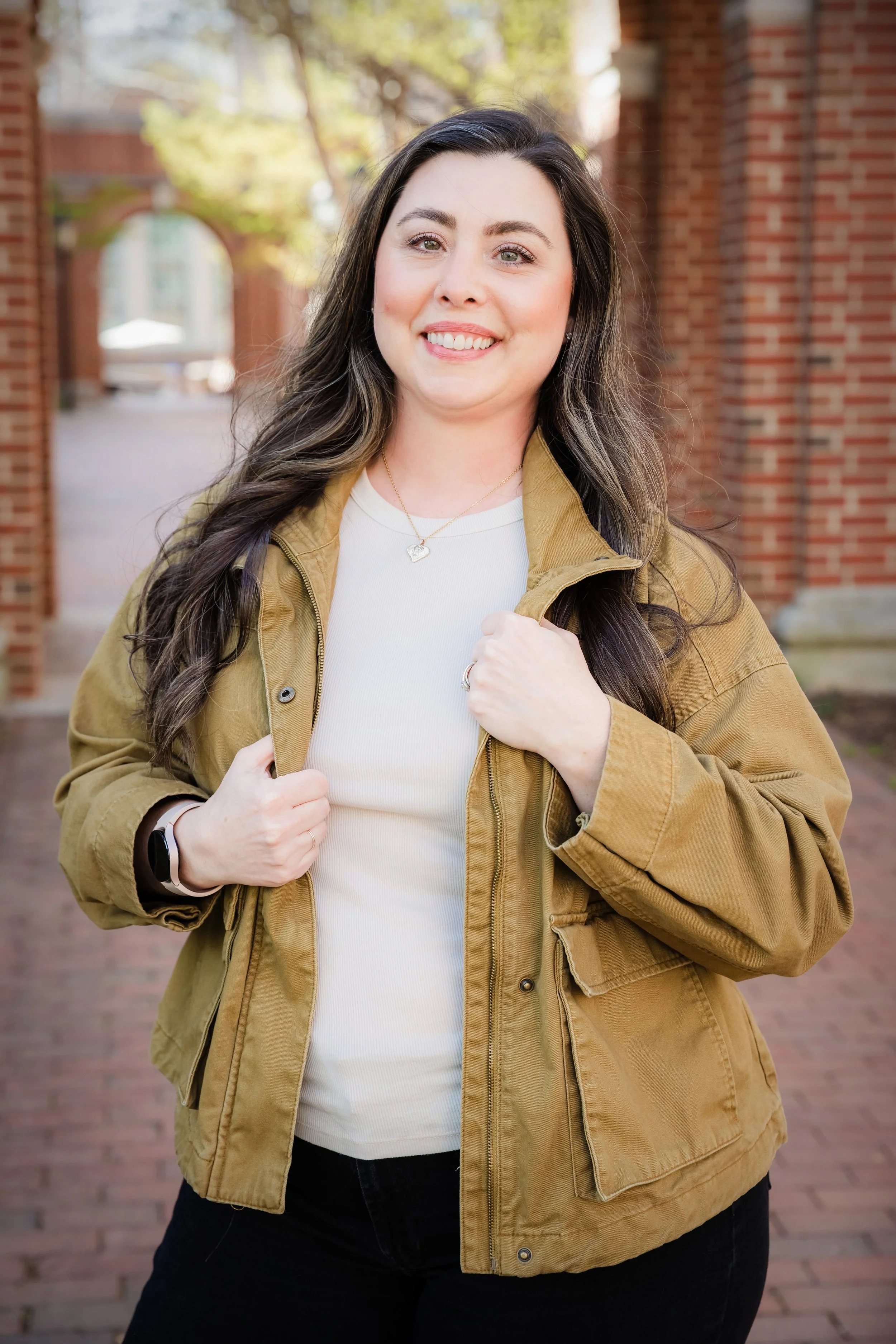A young woman with long dark hair smiling, holding her jacket collar, standing outdoors in front of brick archways and greenery.