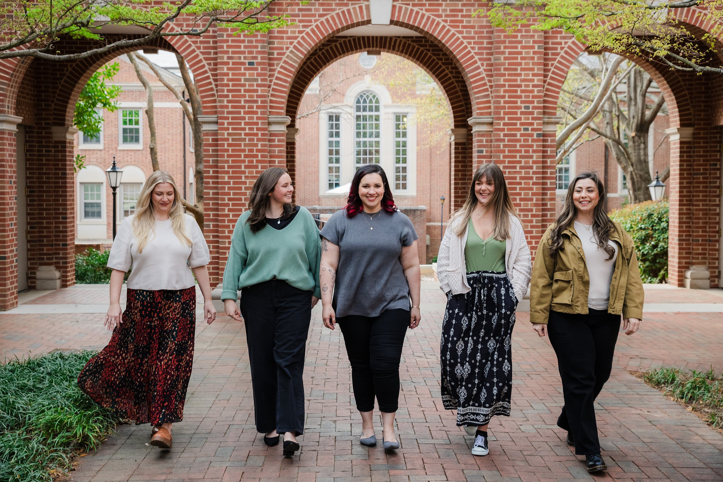 A group of five women walking and smiling outdoors on a brick pathway under trees with brick buildings in the background.