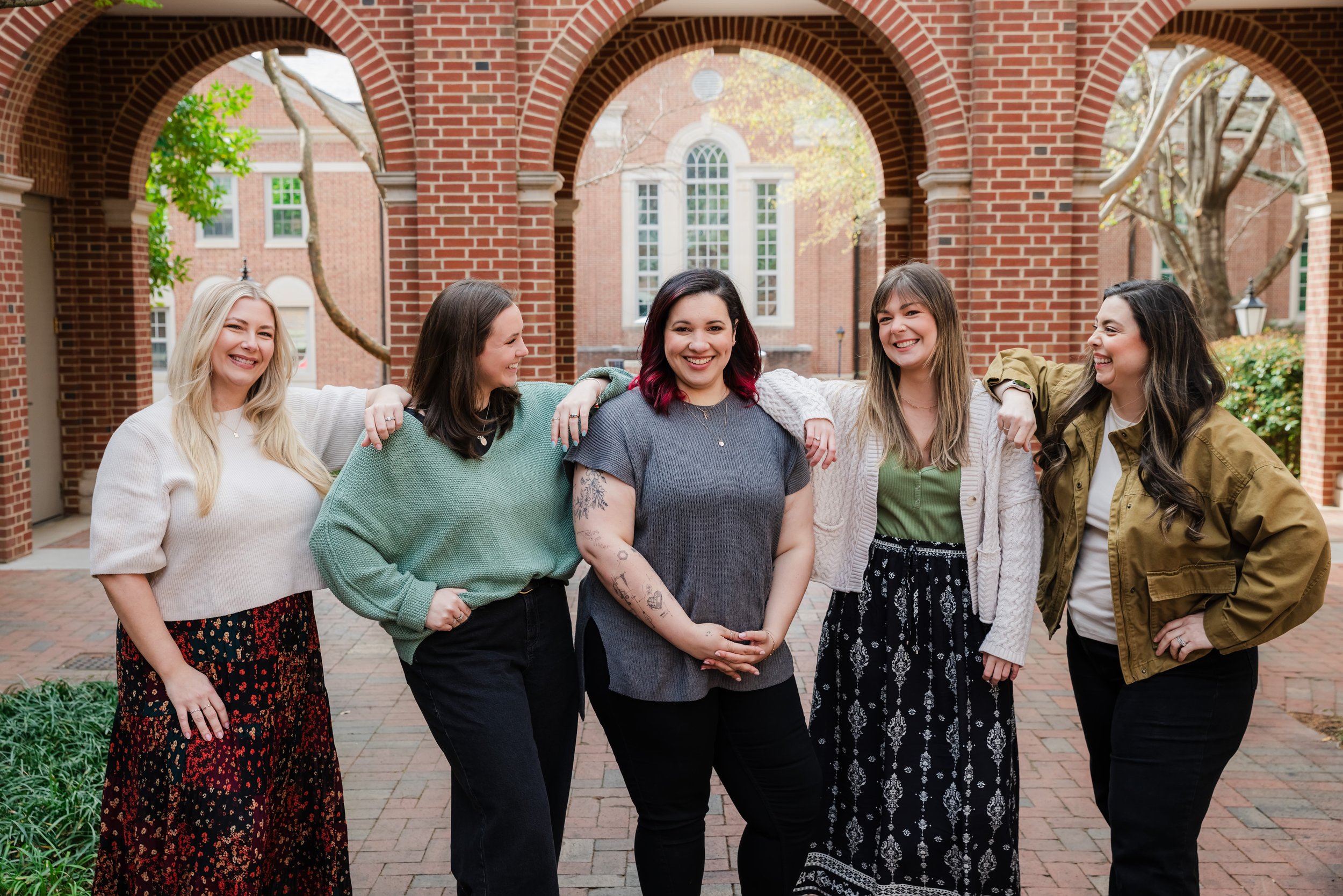 Five women standing close together outdoors, smiling and laughing, with brick arches and a university building in the background.