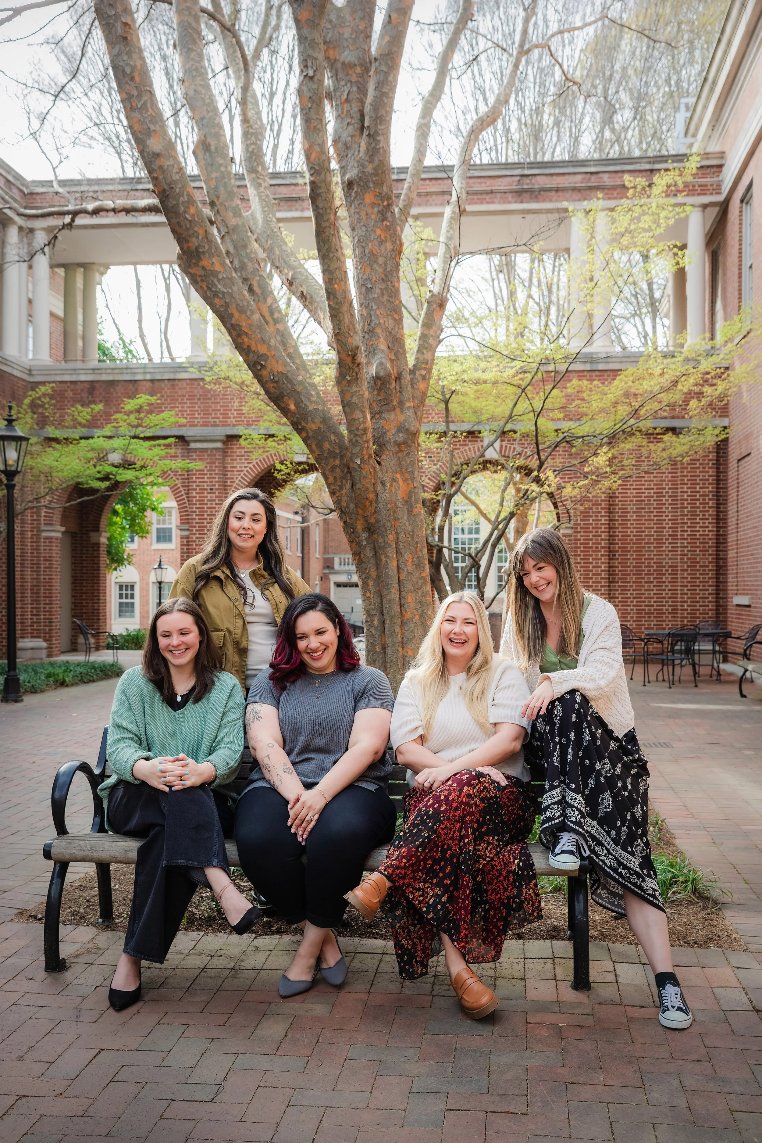 Five women mental health therapists smiling and sitting on a bench in a park with university buildings and trees in the background.
