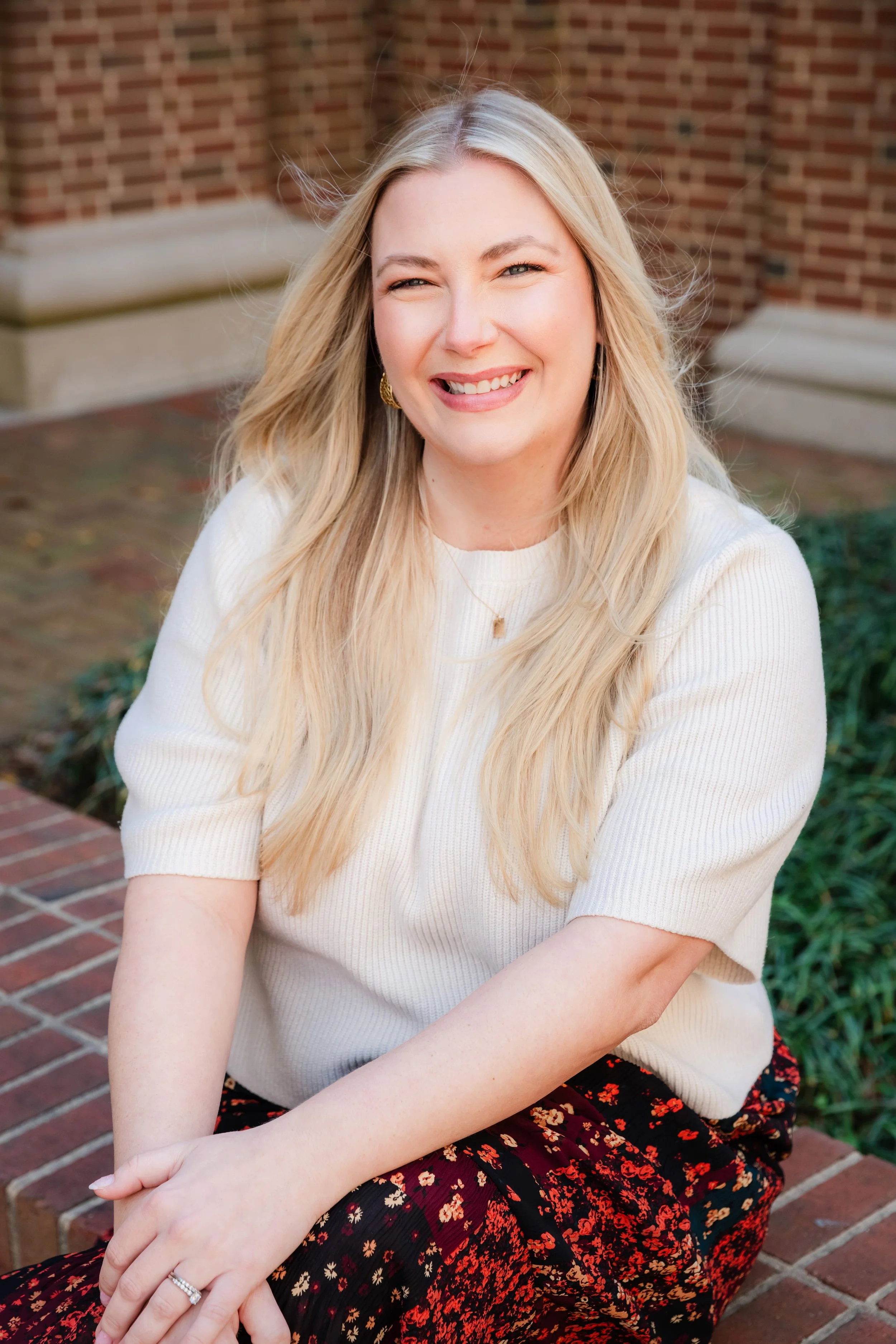 A smiling woman with long blonde hair, wearing a white top and a floral skirt, sitting outdoors near a brick building.