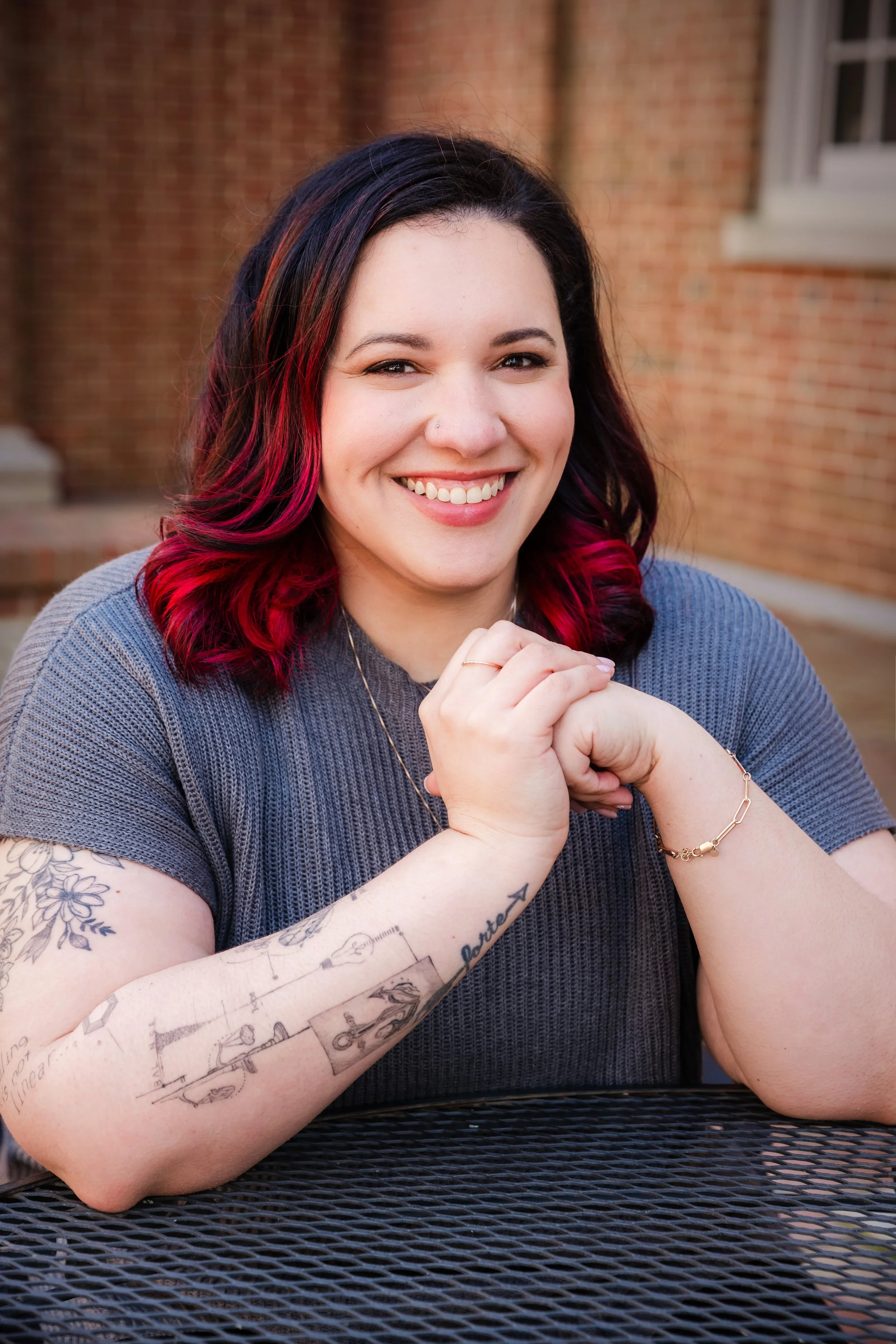 A smiling woman with dark hair with red highlights, sitting at a black metal table outdoors, wearing a gray top, jewelry, and showcasing tattoos on her left arm with a brick building in the background.