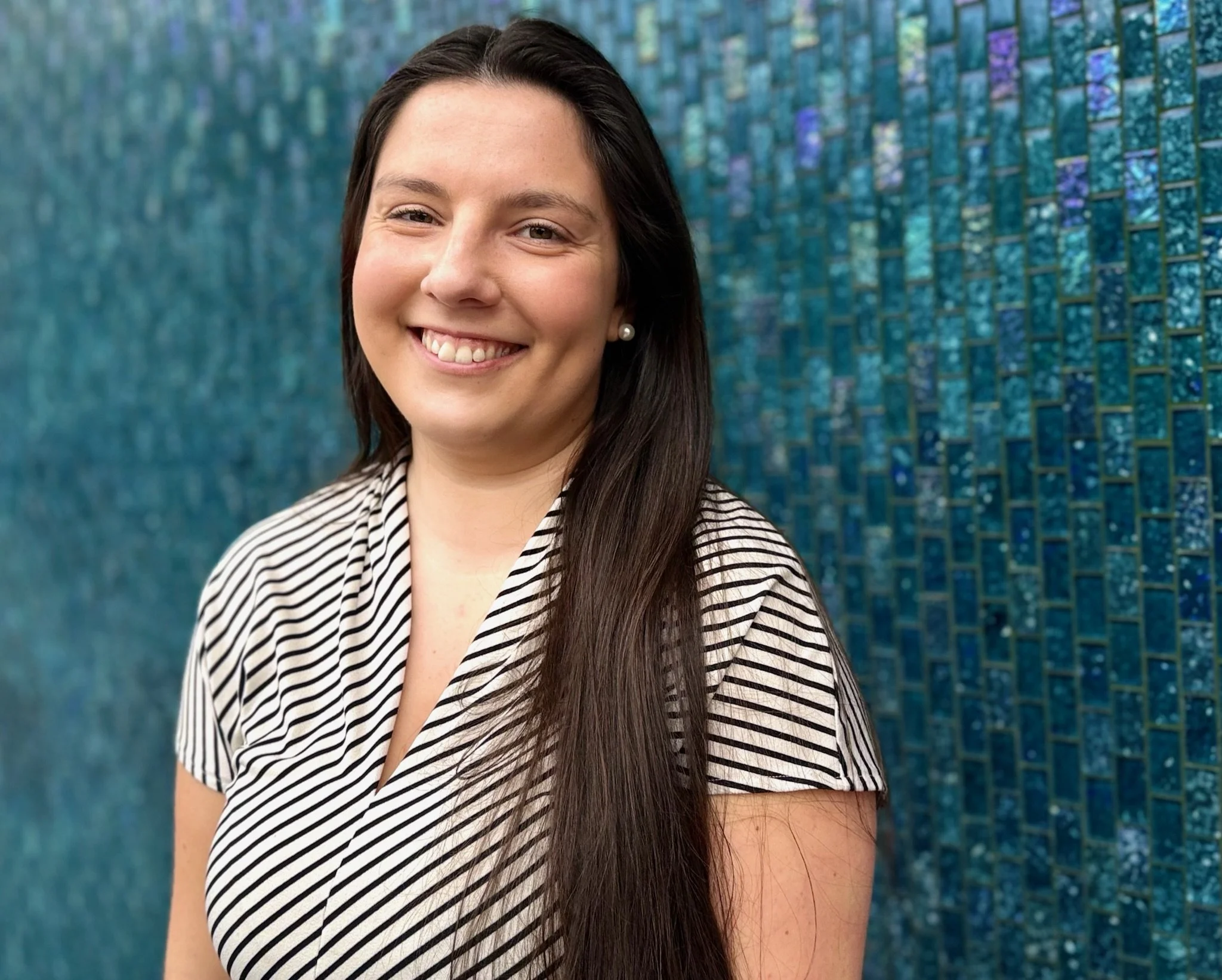 A woman with long dark hair smiling, standing in front of a blue mosaic wall.